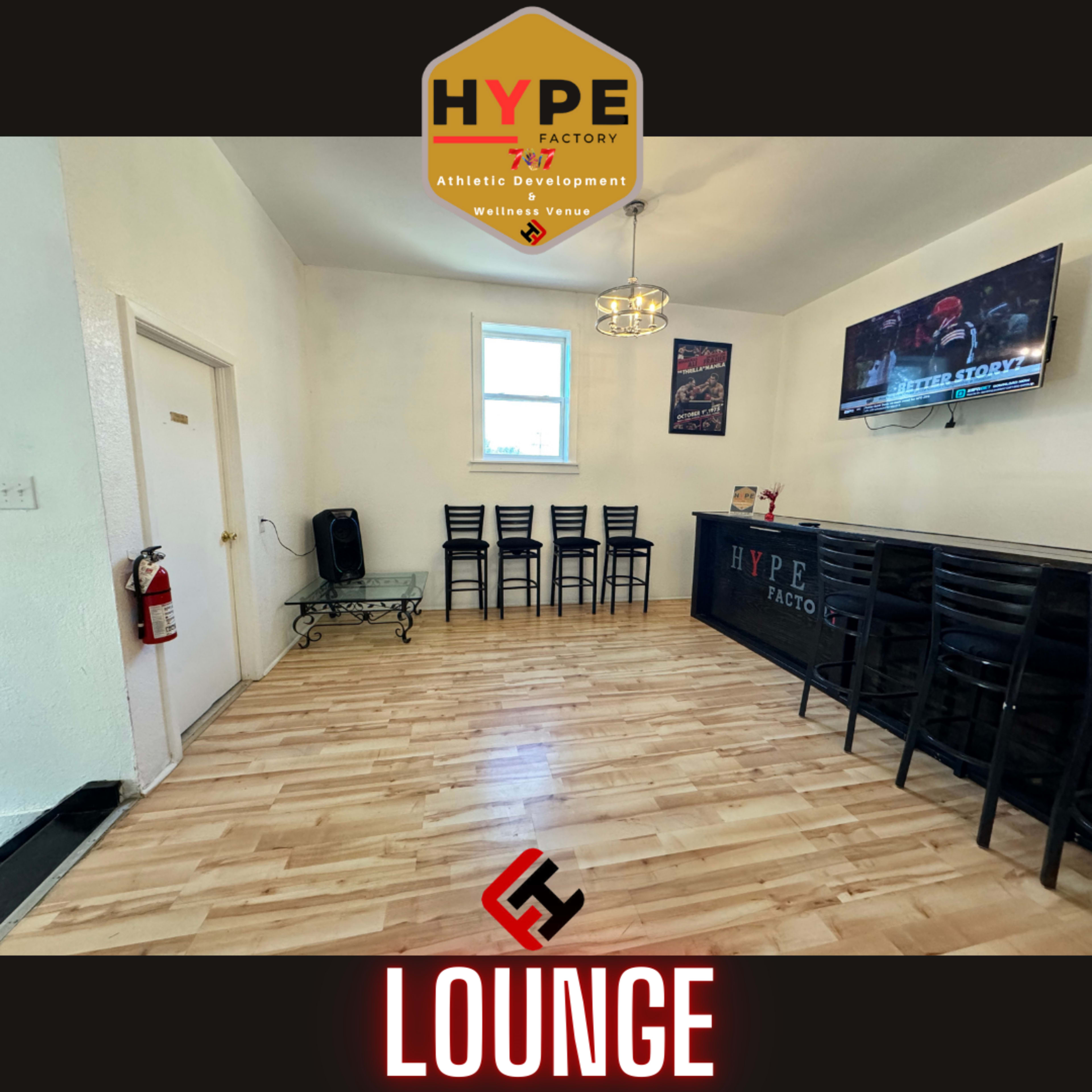 A bright, minimalist lounge area with wooden flooring, featuring a few black chairs, a wall-mounted TV, and a reception area labeled "HYPE FACTORY."