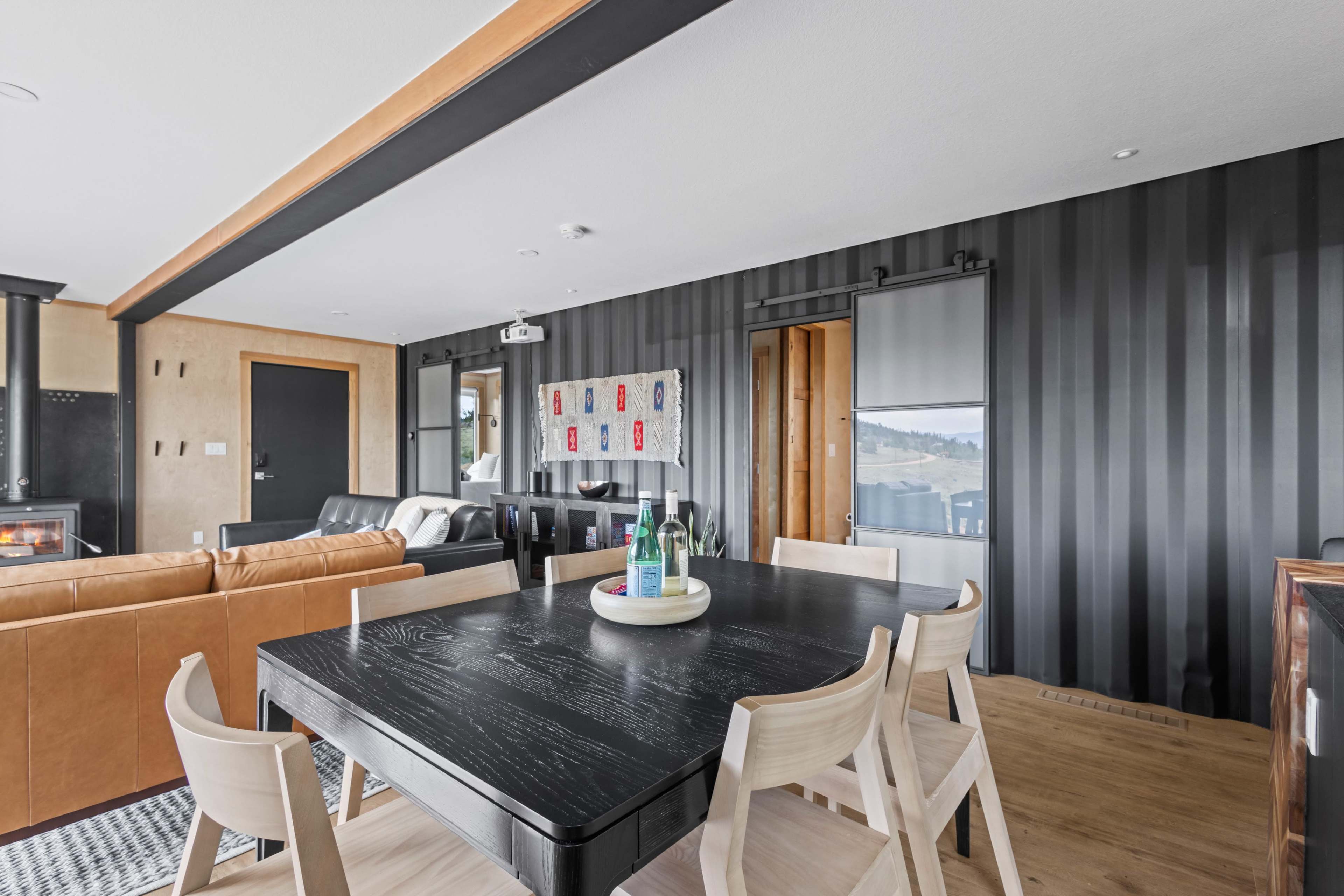 The image shows a modern dining area with a black table surrounded by light wooden chairs, with a living space featuring a leather sofa and a fireplace visible in the background.