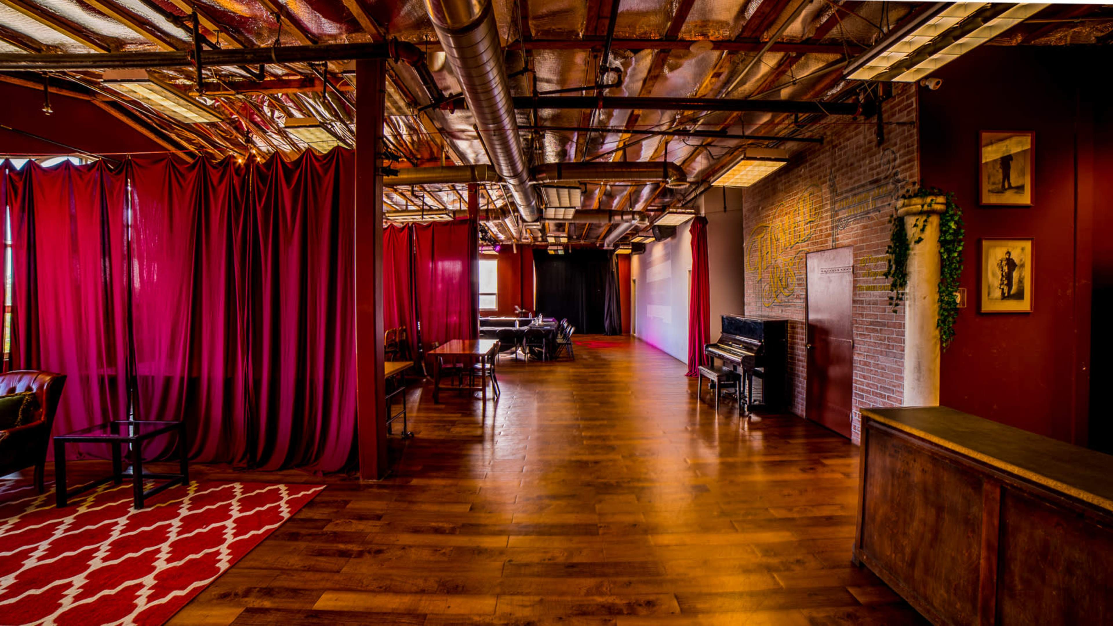 An interior space with wooden floors, exposed beams, a curtain partition in red, and a vintage piano against a brick wall.