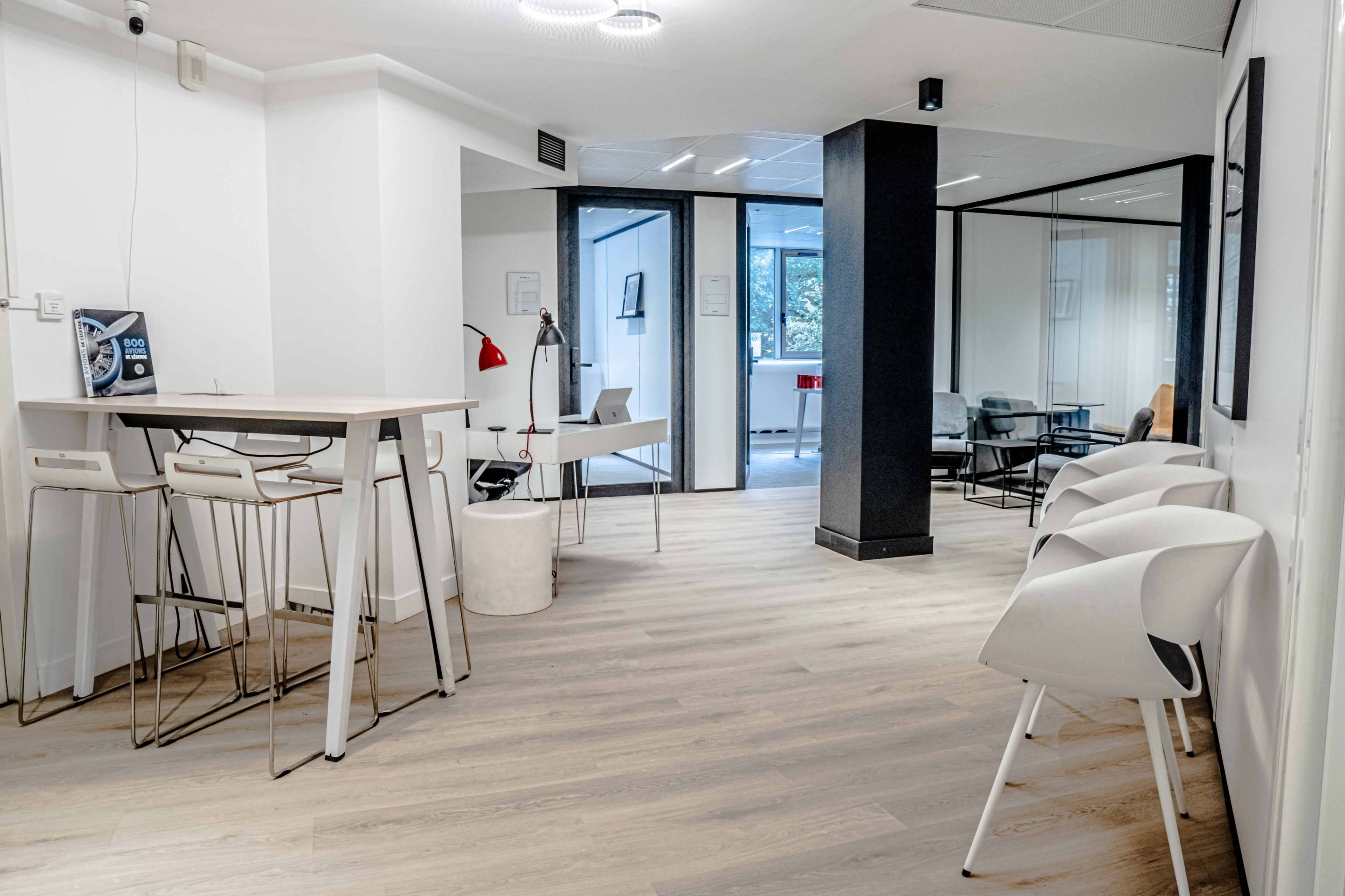 A modern office space featuring a seating area with bar stools, a small table, and glass-walled meeting rooms in the background.