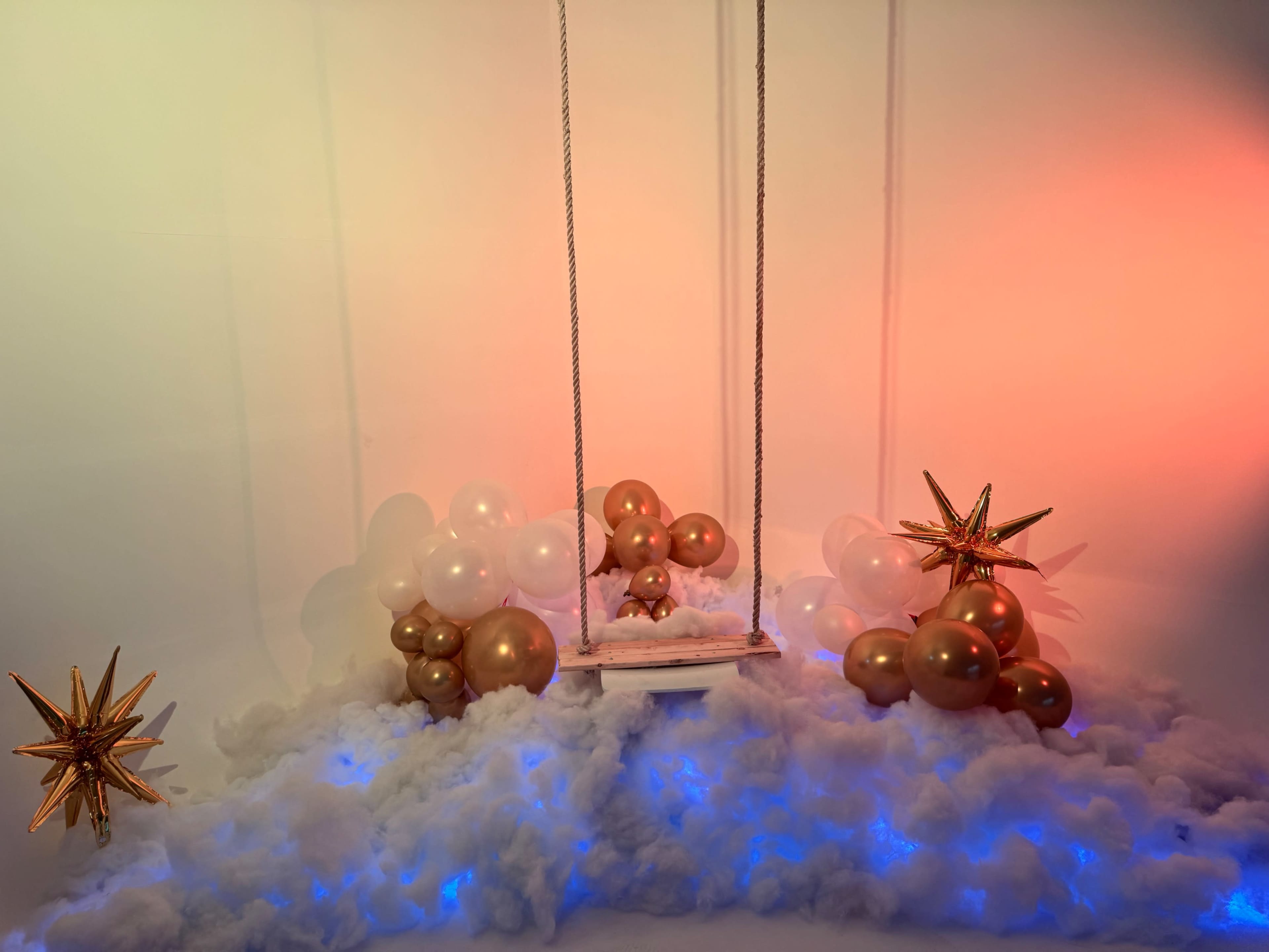 A swing hangs in front of a backdrop decorated with gold balloons and star shapes, resting on a bed of white clouds with blue lighting.
