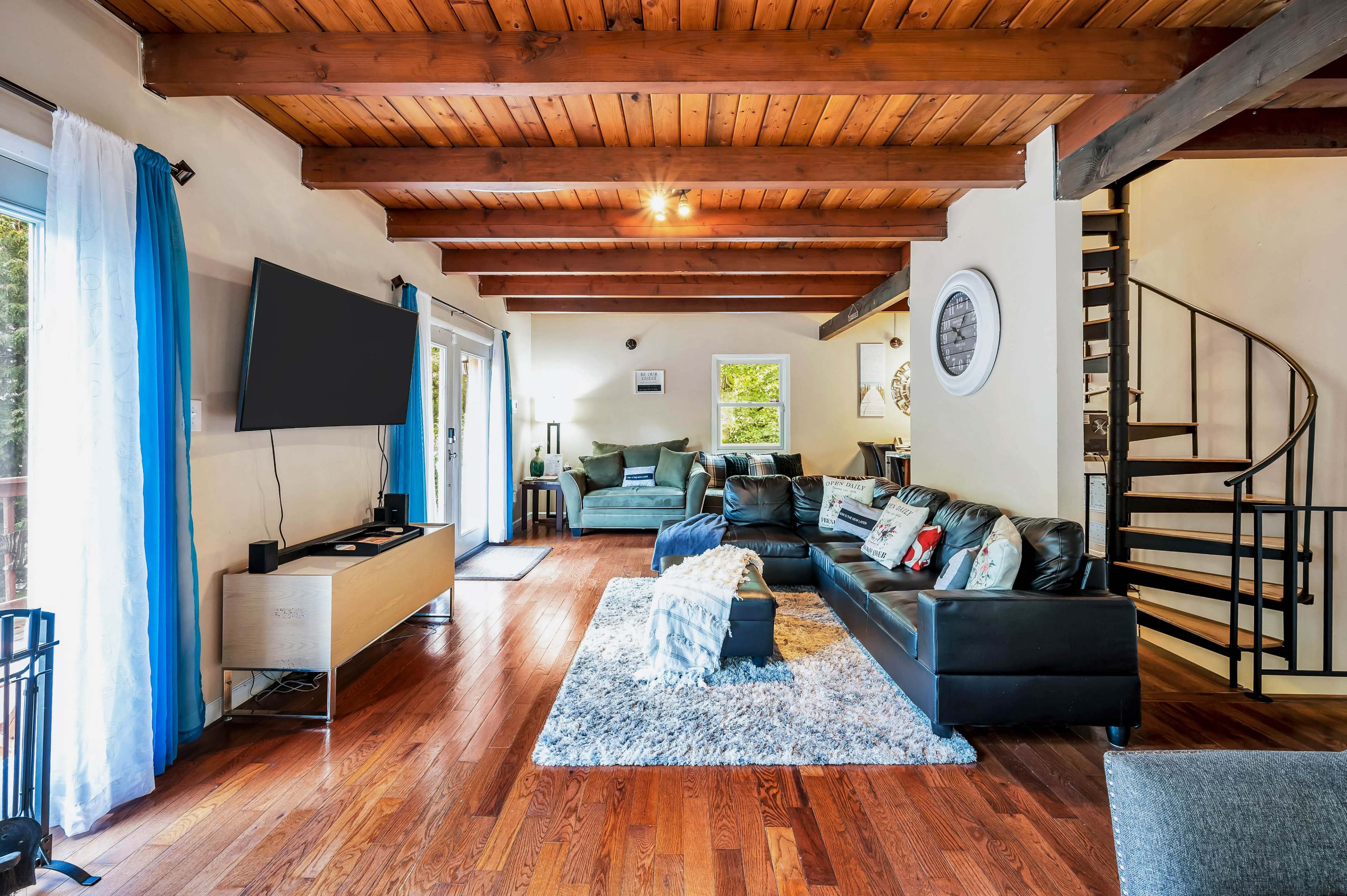 The image shows a spacious living room with wooden beams, leather sofas arranged around a central area rug, and a spiral staircase leading to an upper level.