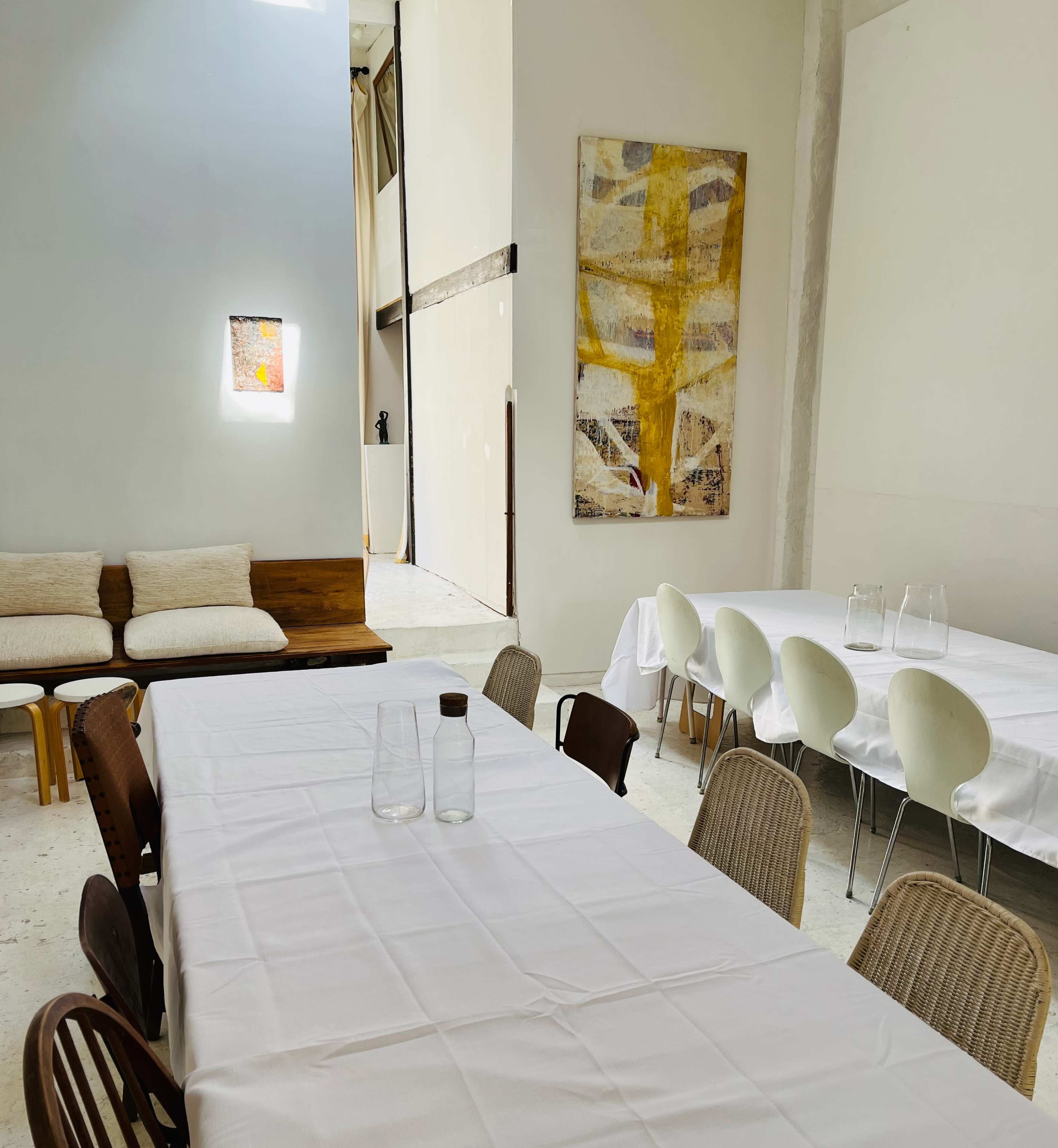 A dining space features a long white table set with glassware, surrounded by various chairs, alongside a minimalist seating area and an abstract artwork on the wall.