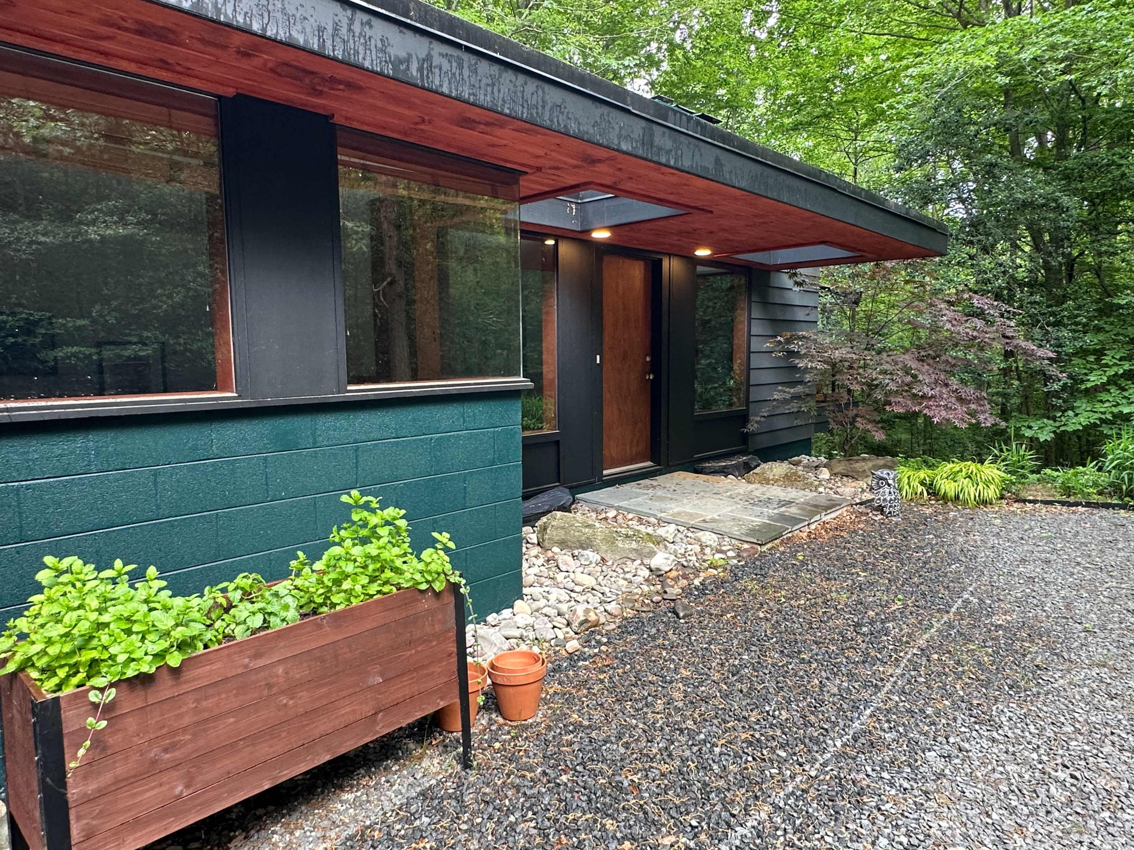 A modern house with a sloped roof features a wooden door, large windows, and a gravel pathway surrounded by greenery.