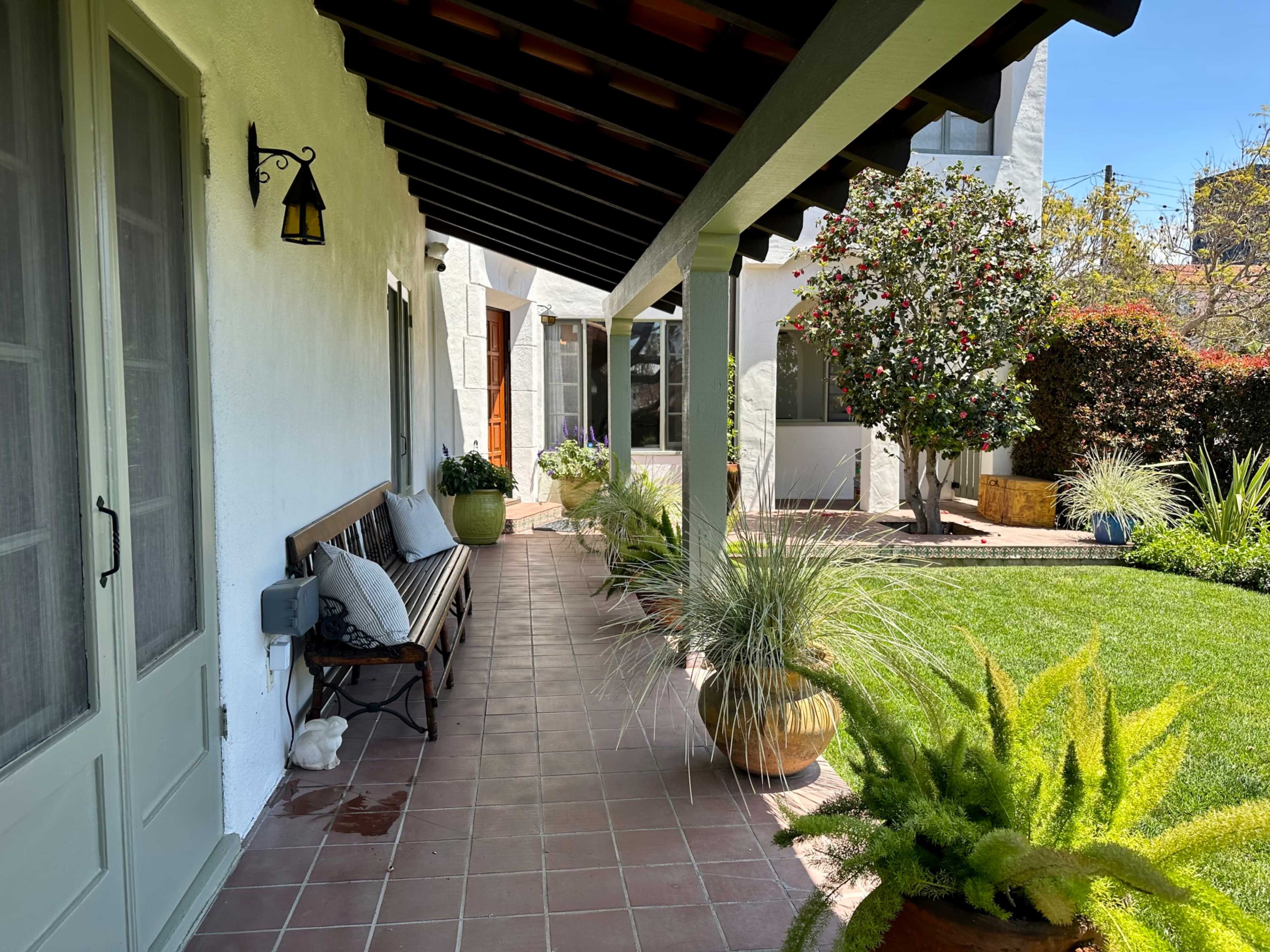 A landscaped walkway beside a house, featuring potted plants and a bench under a covered porch.