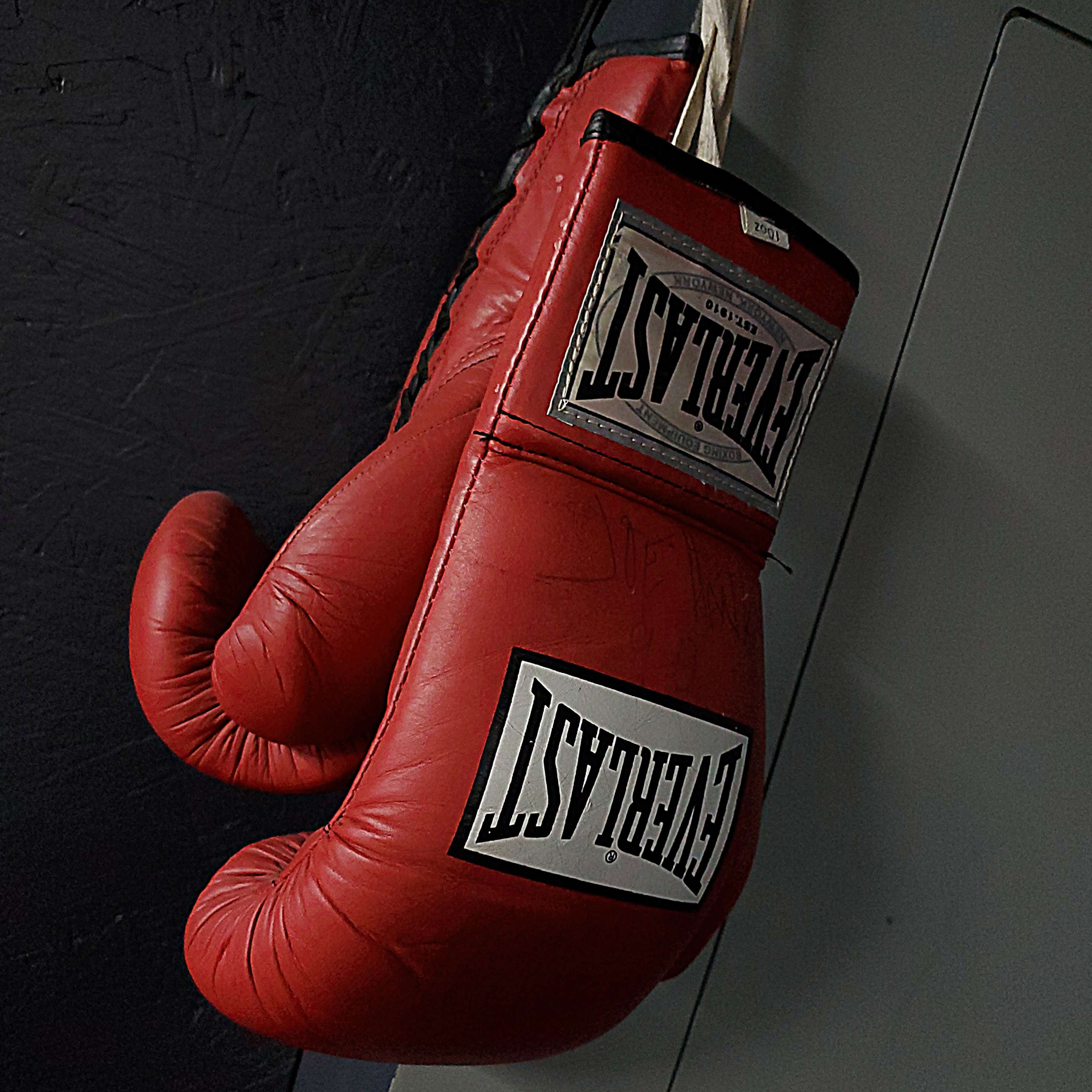 A pair of red boxing gloves is hanging at an angle from a hook against a dark wall.