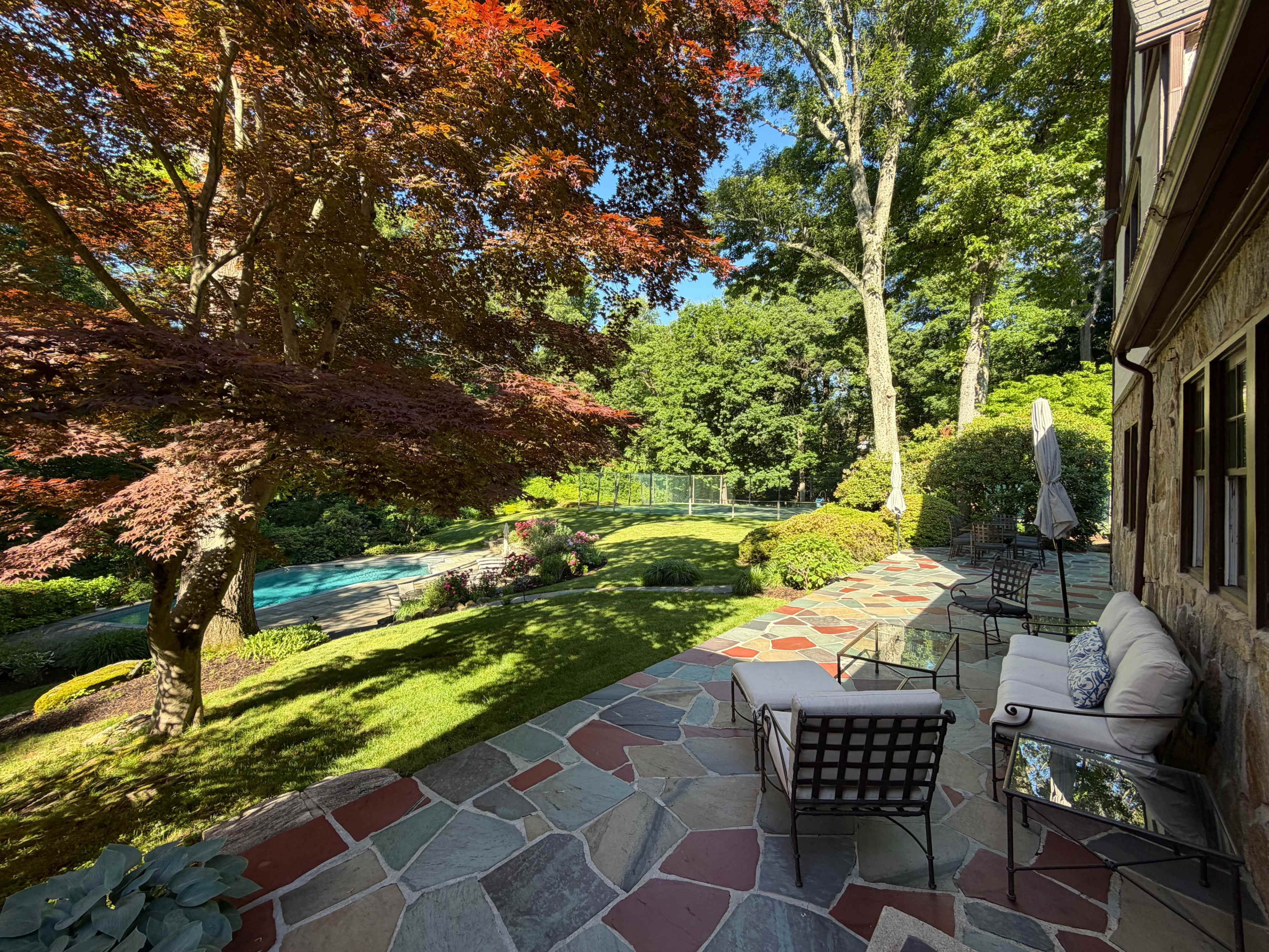 The image shows a landscaped backyard featuring a patio with seating, a swimming pool, and lush greenery, framed by a vibrant tree.