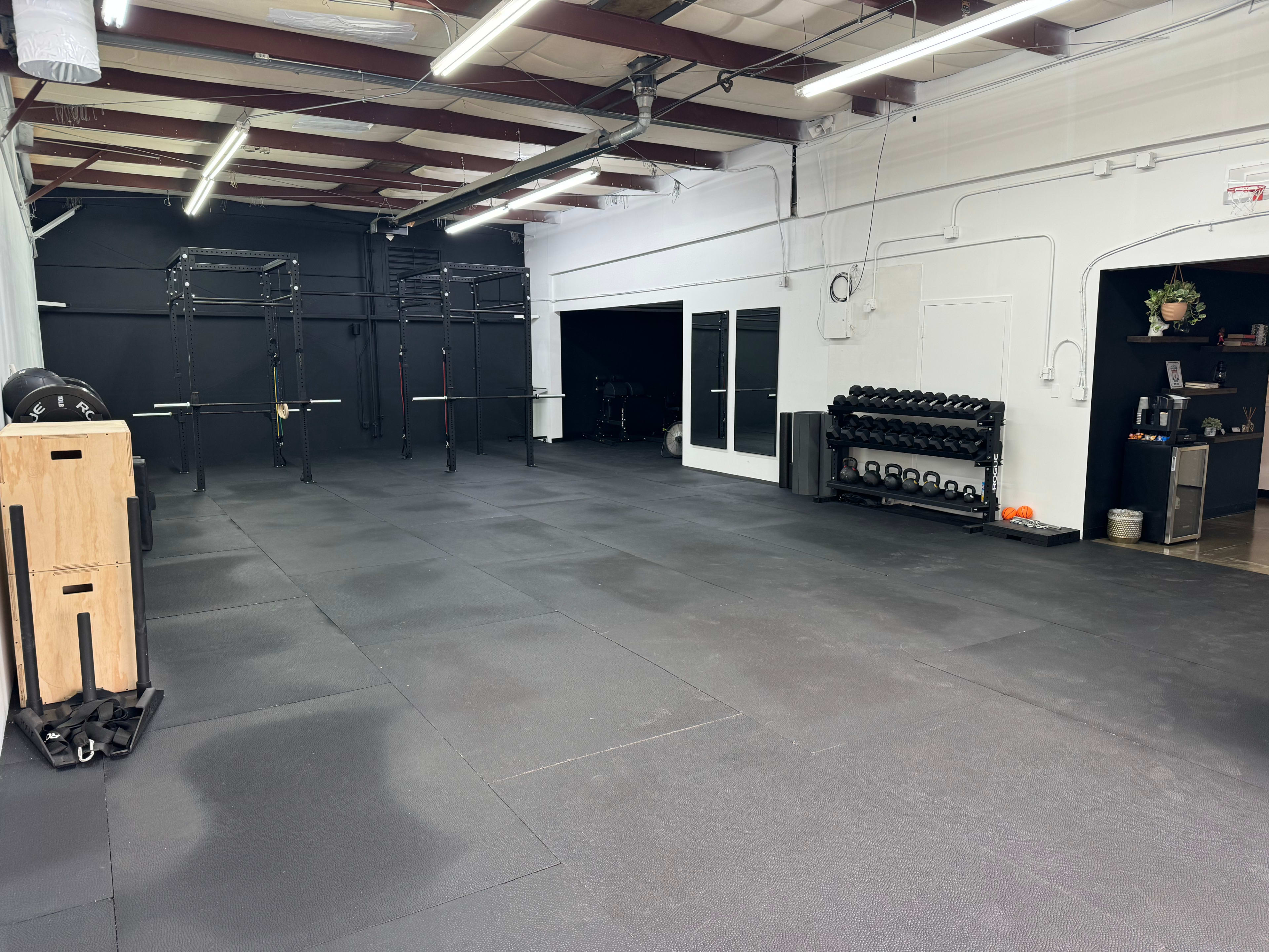 The image shows an empty gym space with rubber flooring, strength training equipment, and a rack of dumbbells against the wall.