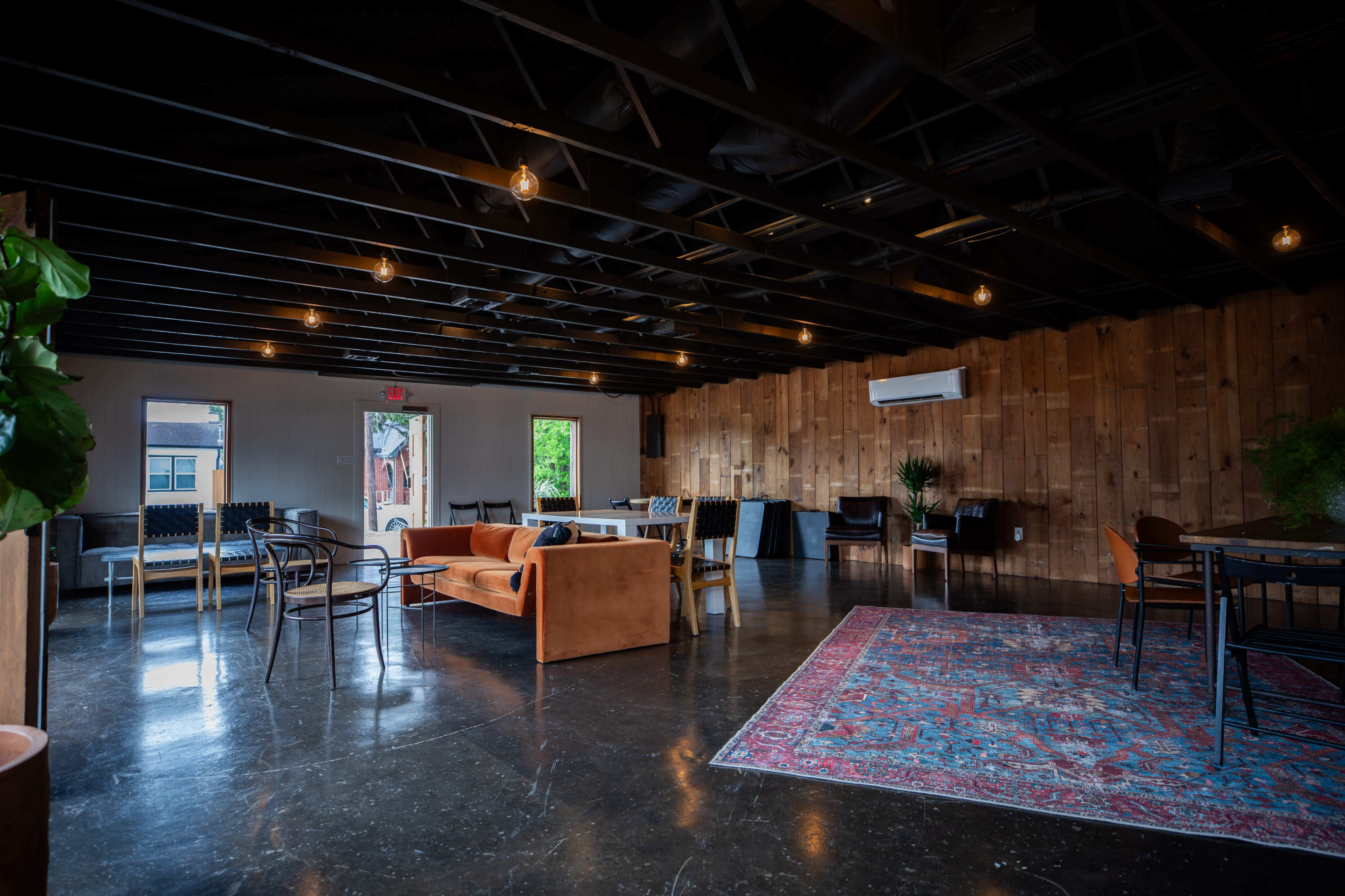 The image shows a spacious interior with a wooden accent wall, various seating options including a brown couch, and a large rug on a polished floor, illuminated by pendant lights.