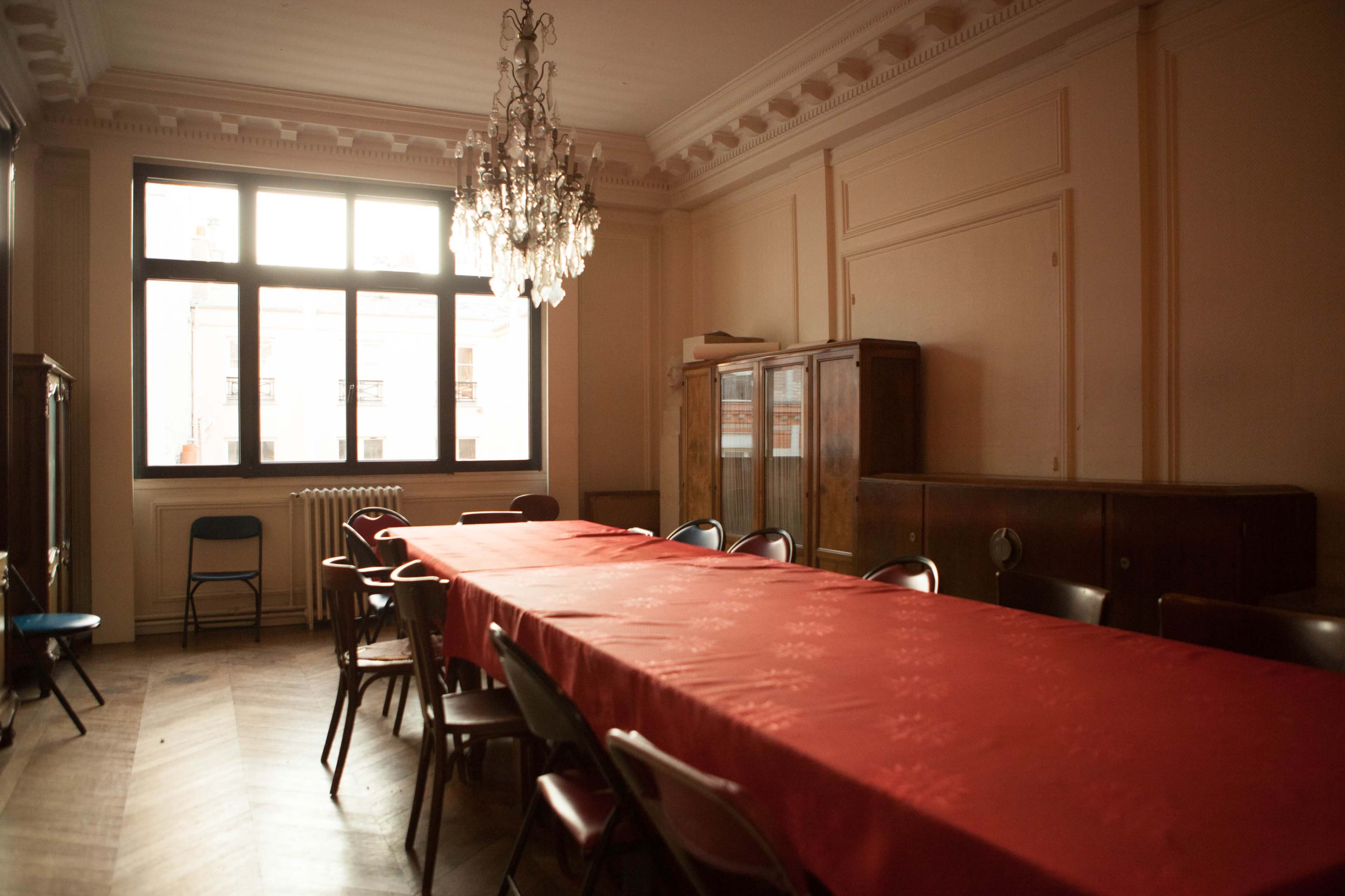 The image features a spacious room with a long red table surrounded by chairs, illuminated by natural light from a large window.