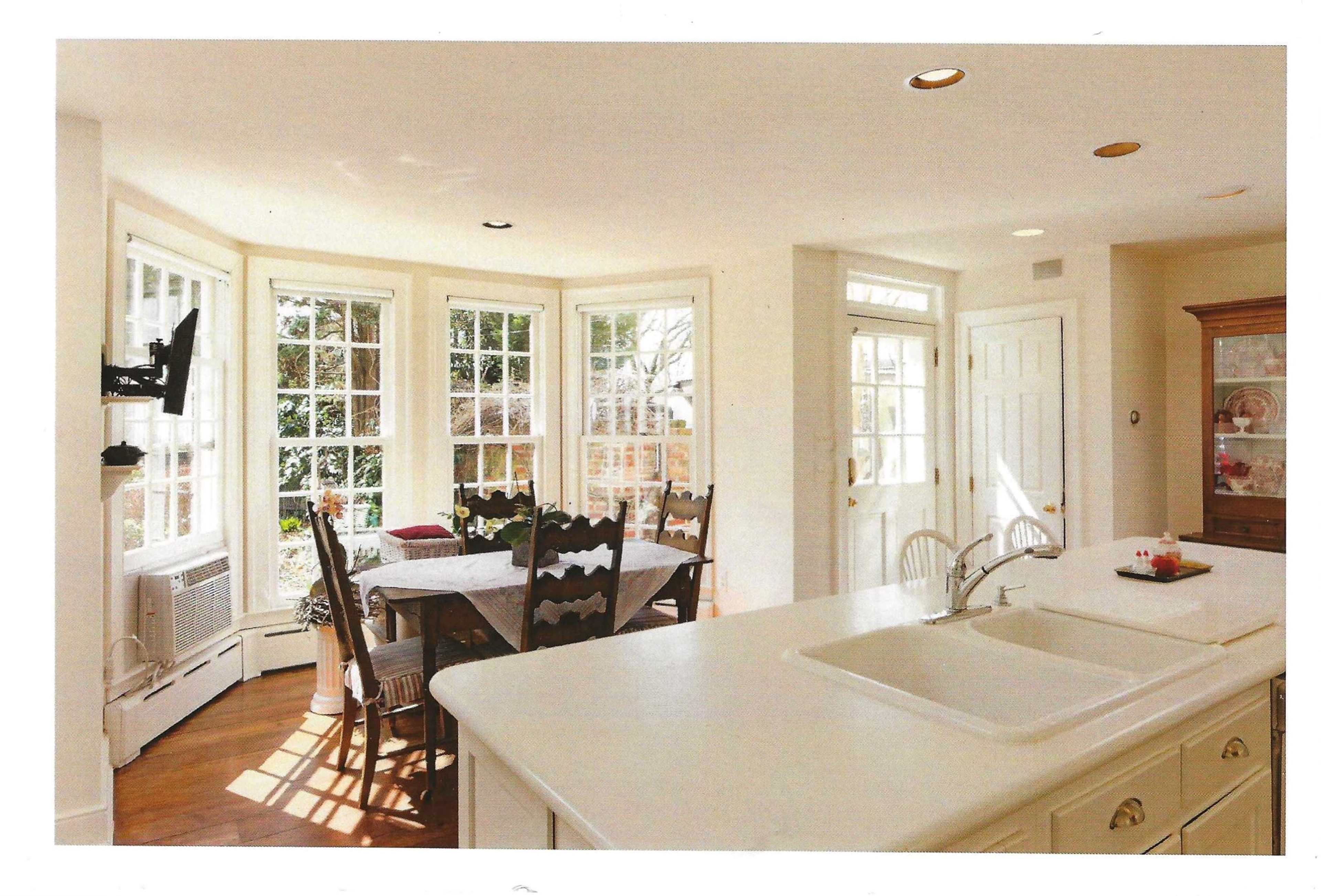 The image shows a bright kitchen with a large windowed dining area, a central island, and a wooden dining table surrounded by chairs.