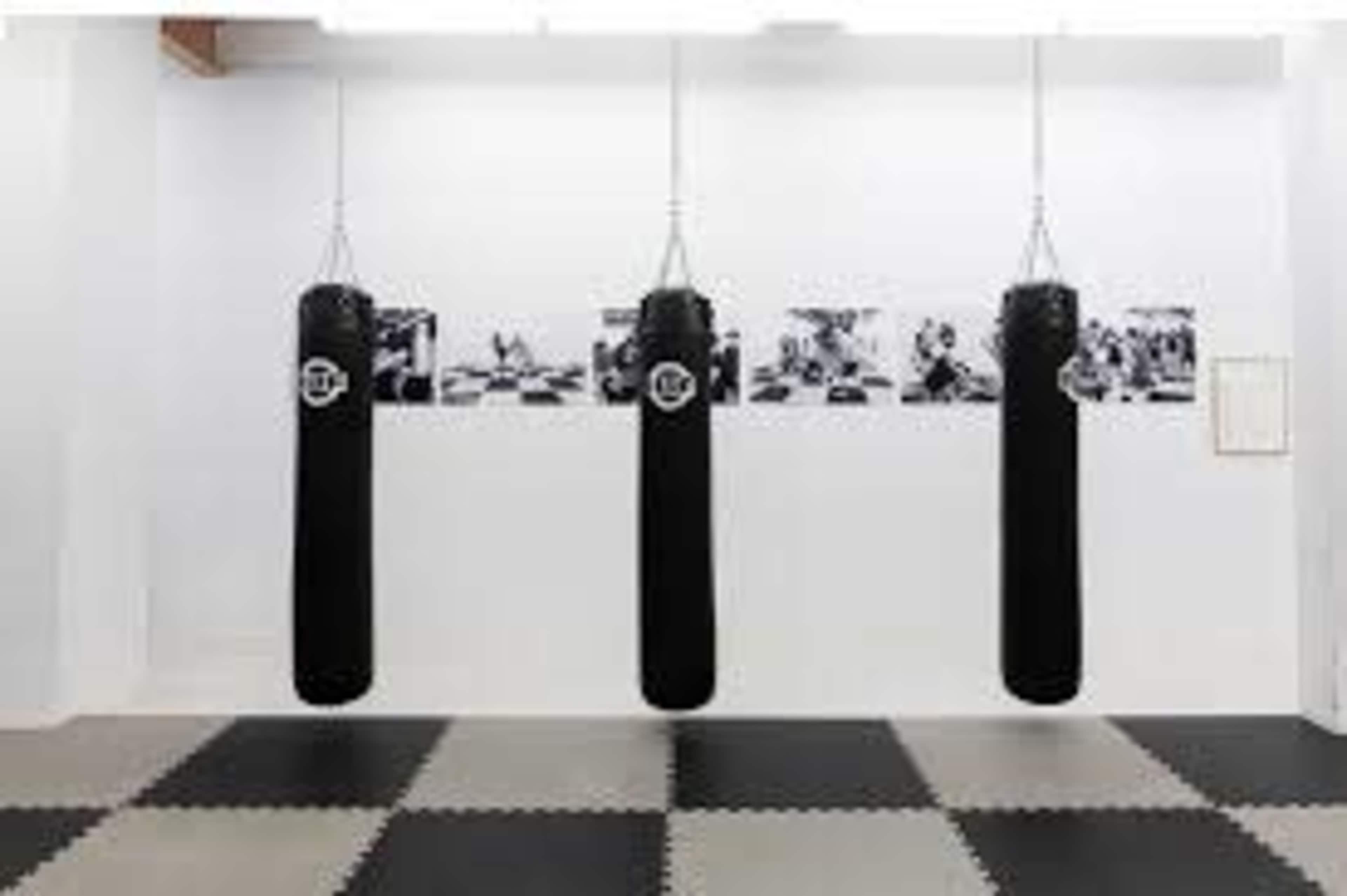 Three black punching bags hanging in a gym with black and white photographs displayed on the wall behind them.