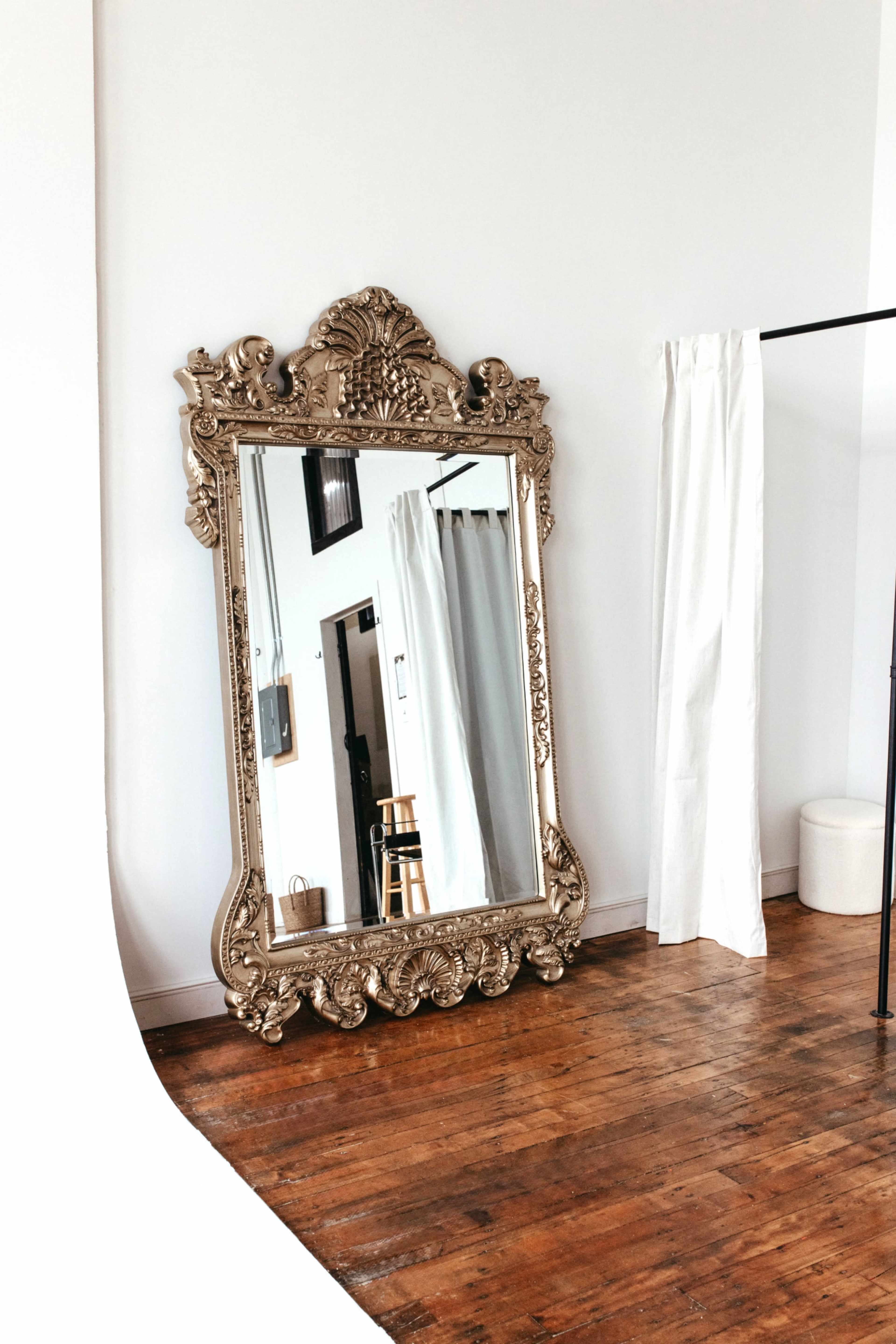A large, ornate mirror with a decorative frame stands against a white wall in a bright, minimalist room featuring wooden floors.