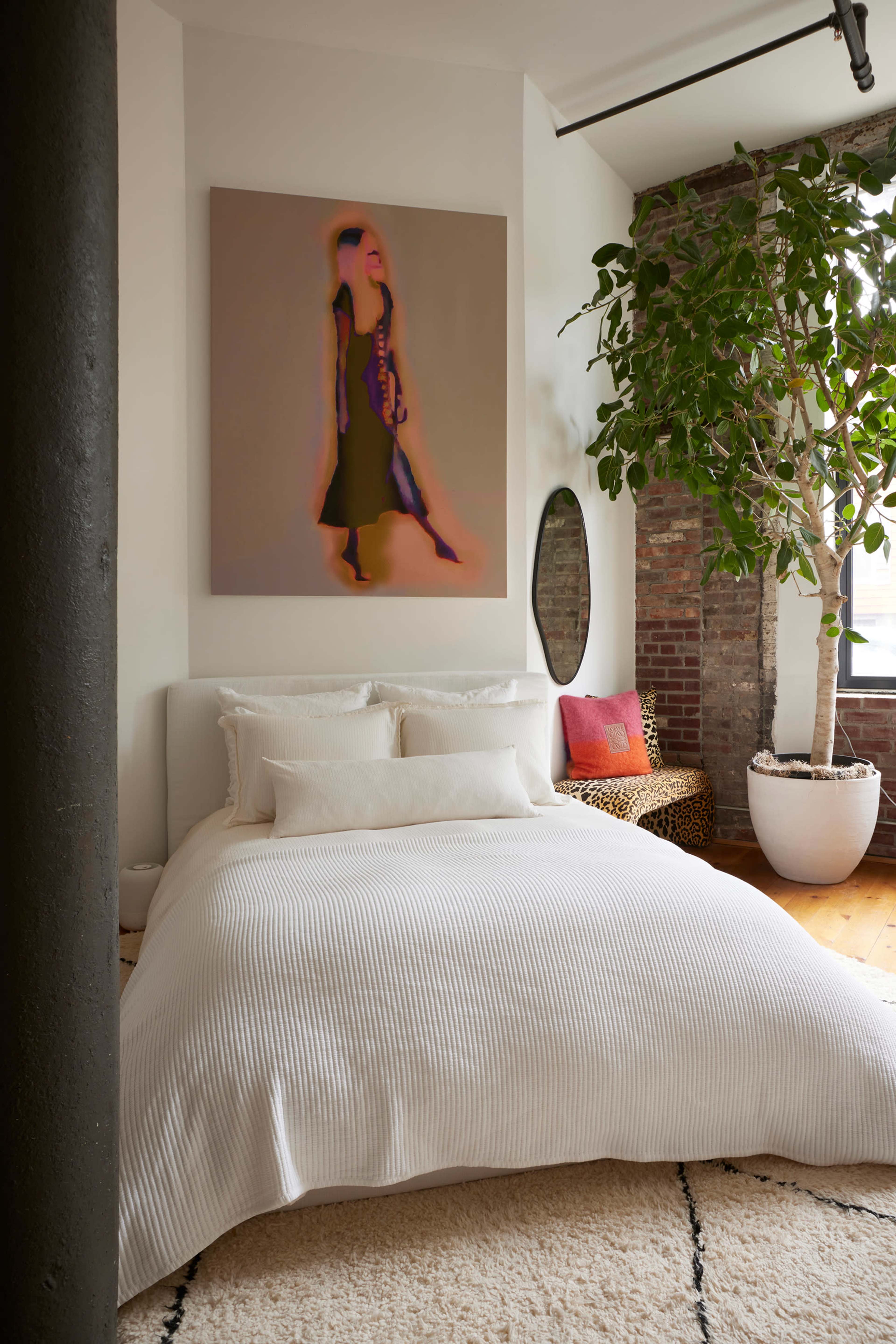 The image shows a modern bedroom with a large bed covered in a white blanket, a prominent abstract artwork on the wall, and a potted plant in the corner.