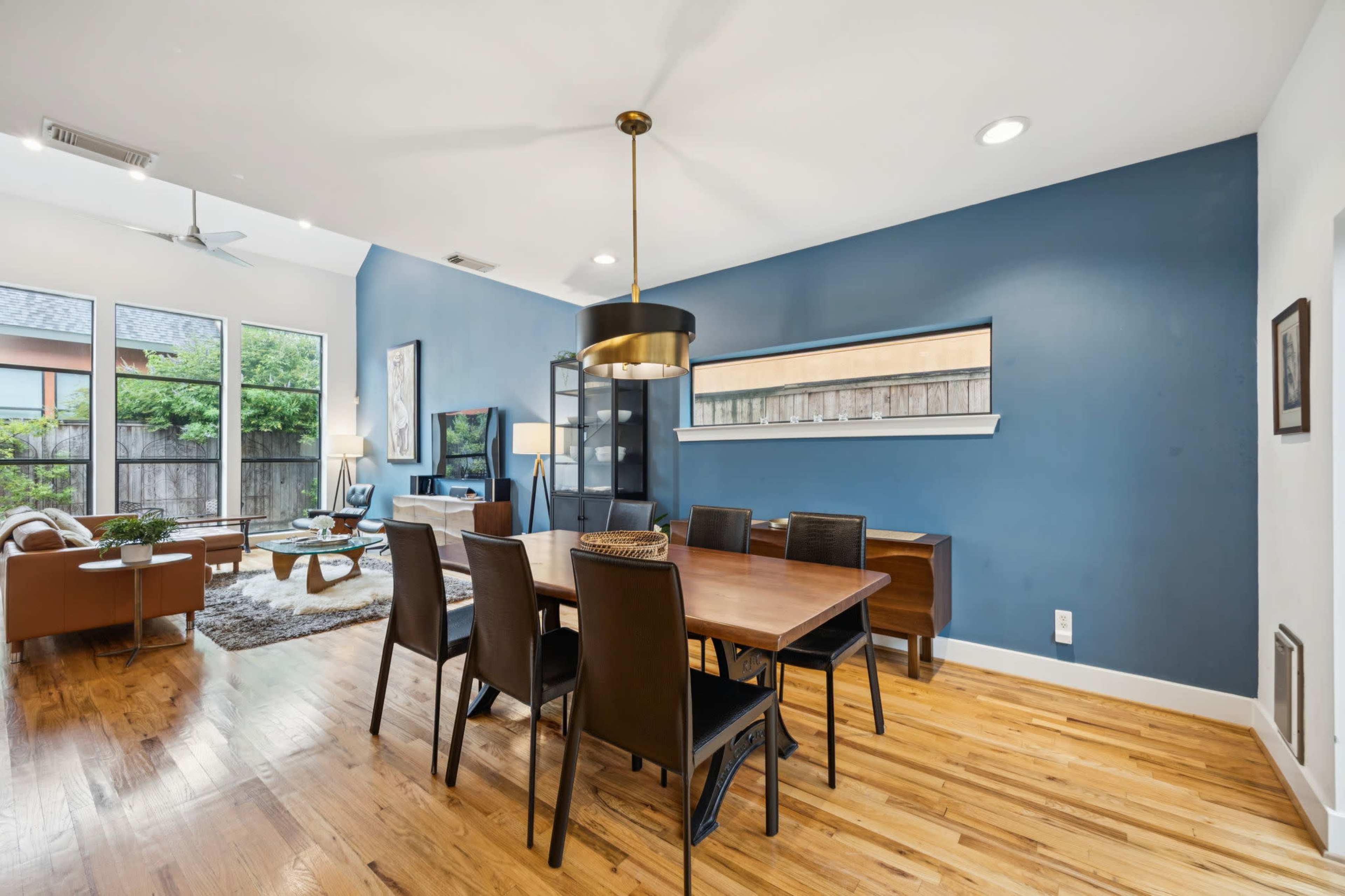 The image shows a modern dining area with a wooden table set for six, adjacent to a living space featuring a television and large windows that provide natural light.