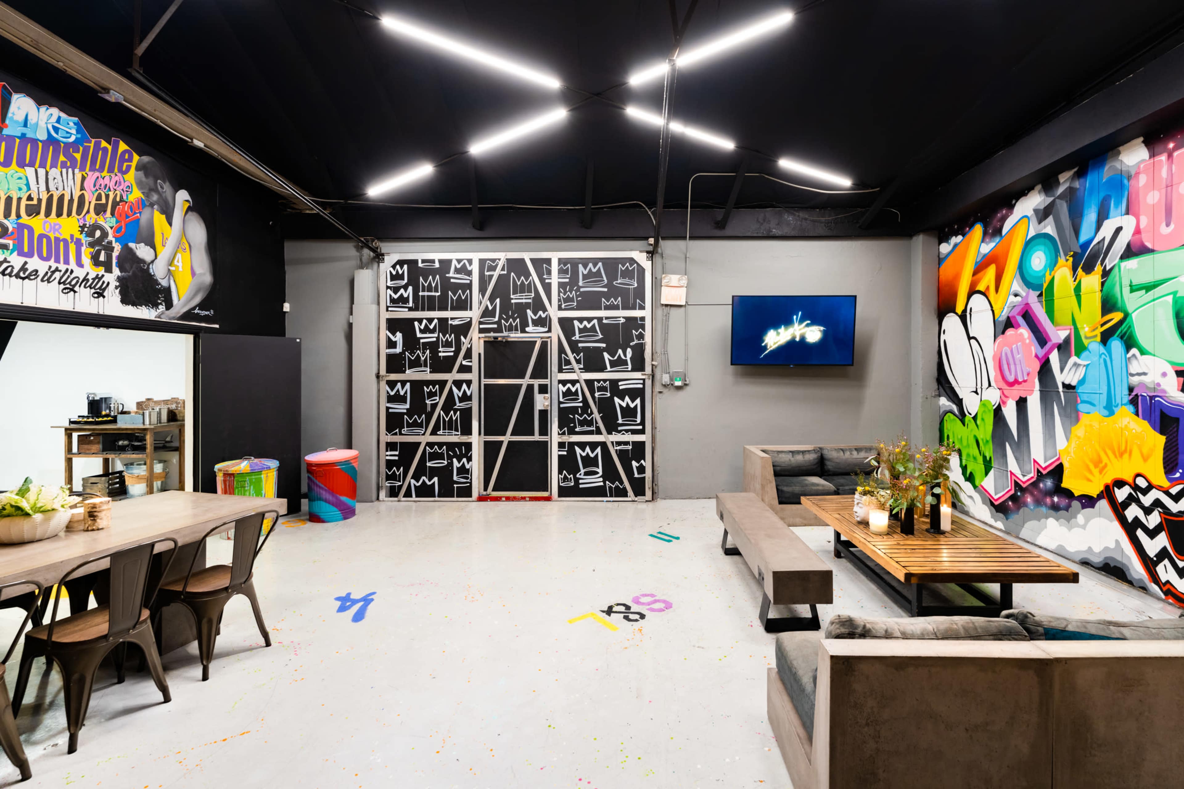 A modern, open space featuring a dining table, a lounge area, a colorful mural on one wall, and a large television mounted on another wall.