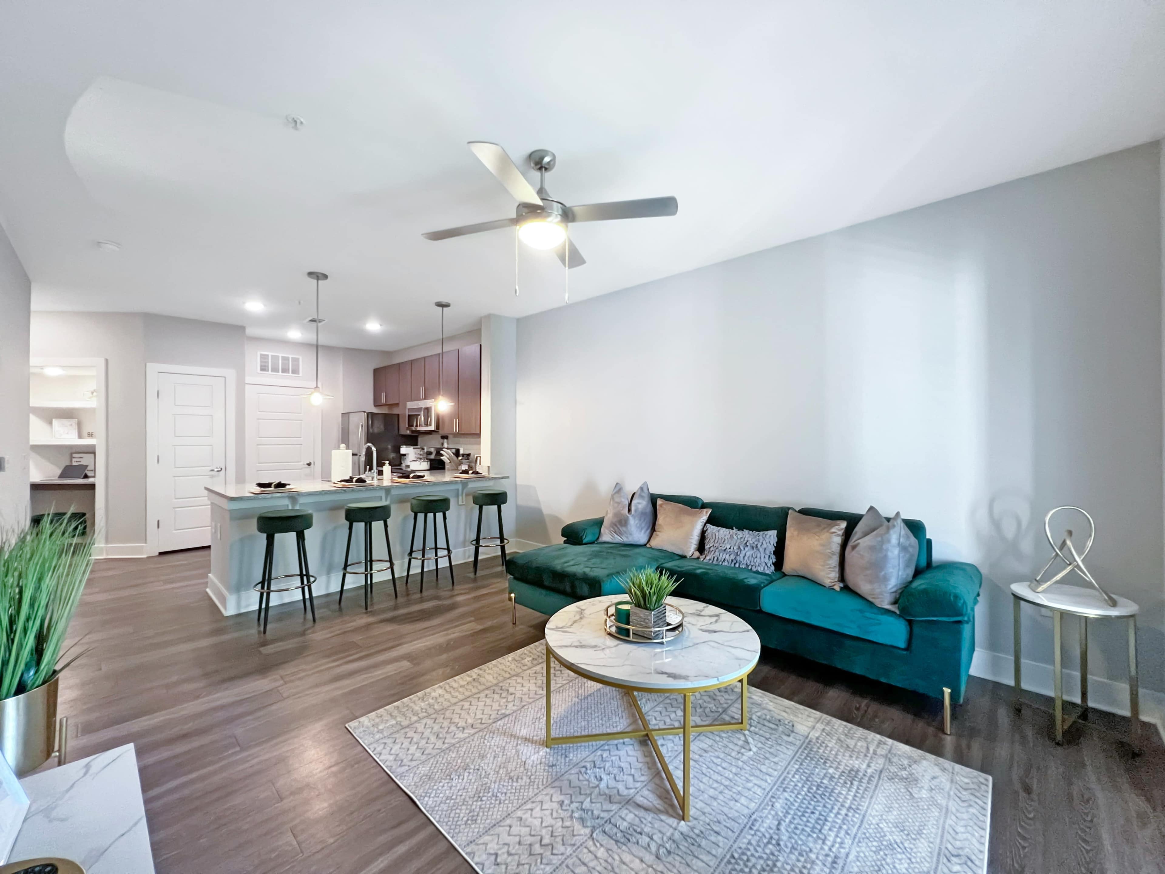 A modern living room with a green sofa, a round coffee table, and a kitchen area featuring bar stools and cabinetry in the background.