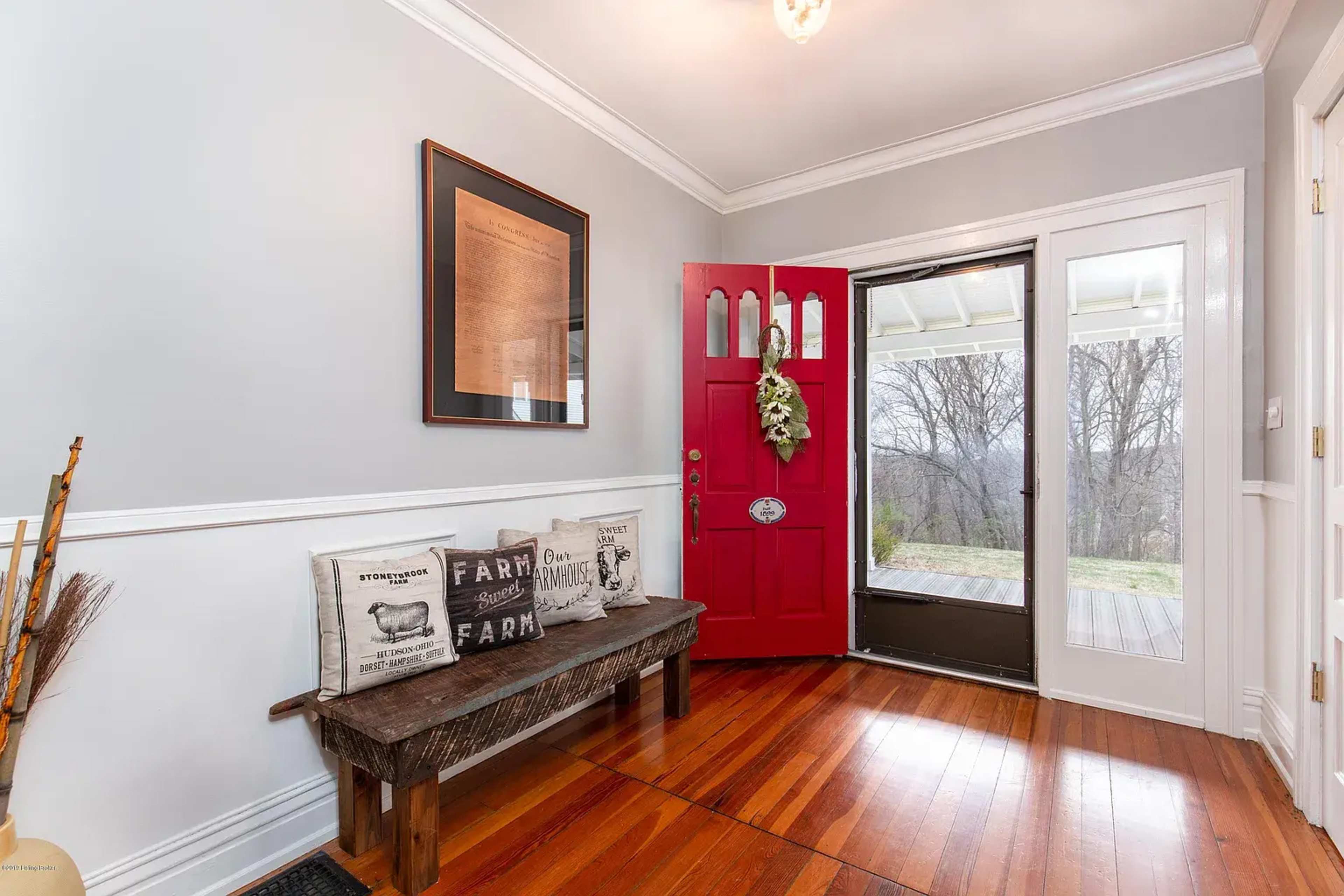The entryway features a red front door, a wooden bench with decorative pillows, and a view of a backyard through a screened door.