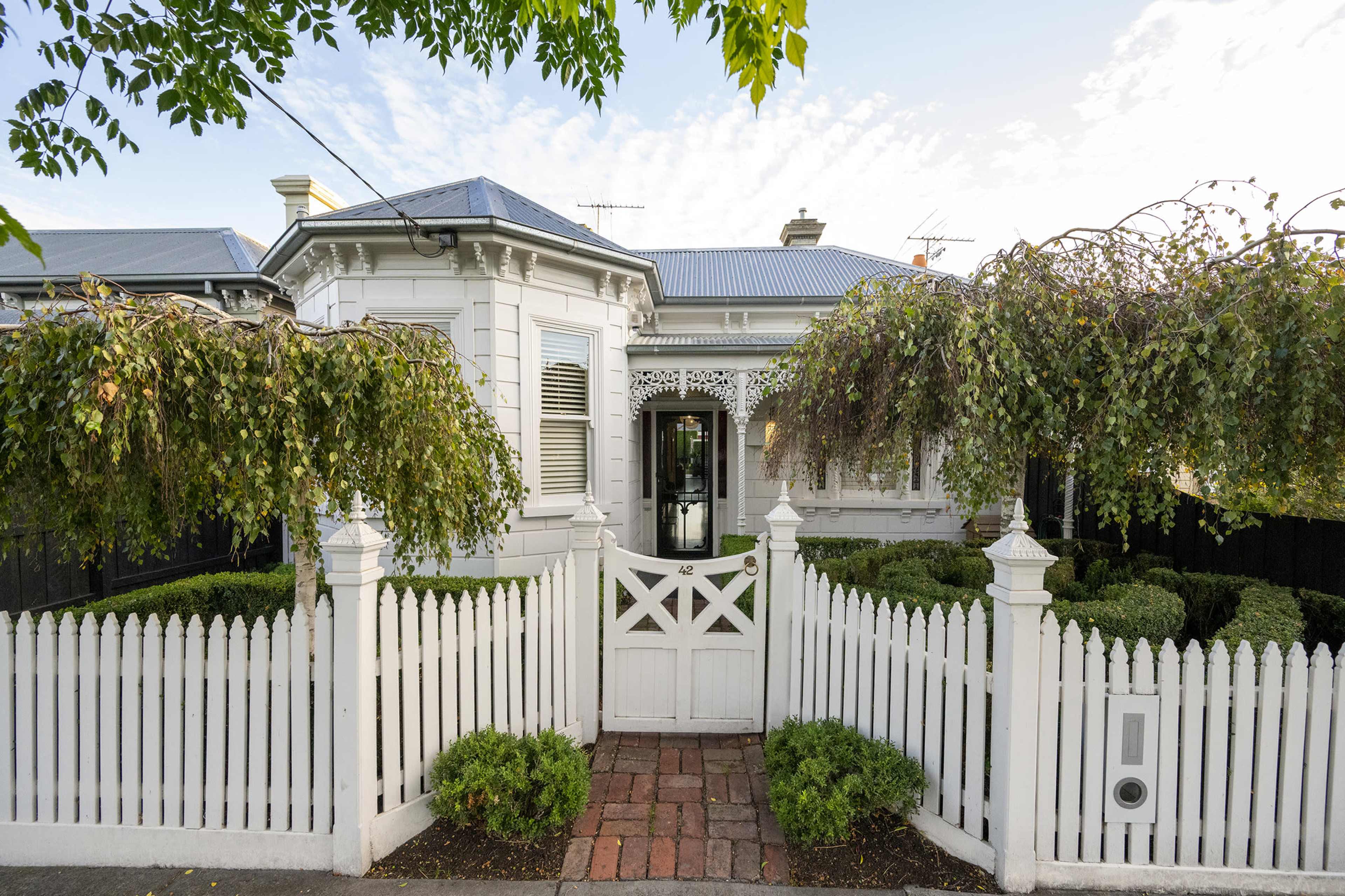 The image shows a white Victorian-style house with a picket fence and well-manicured hedges in the front yard.