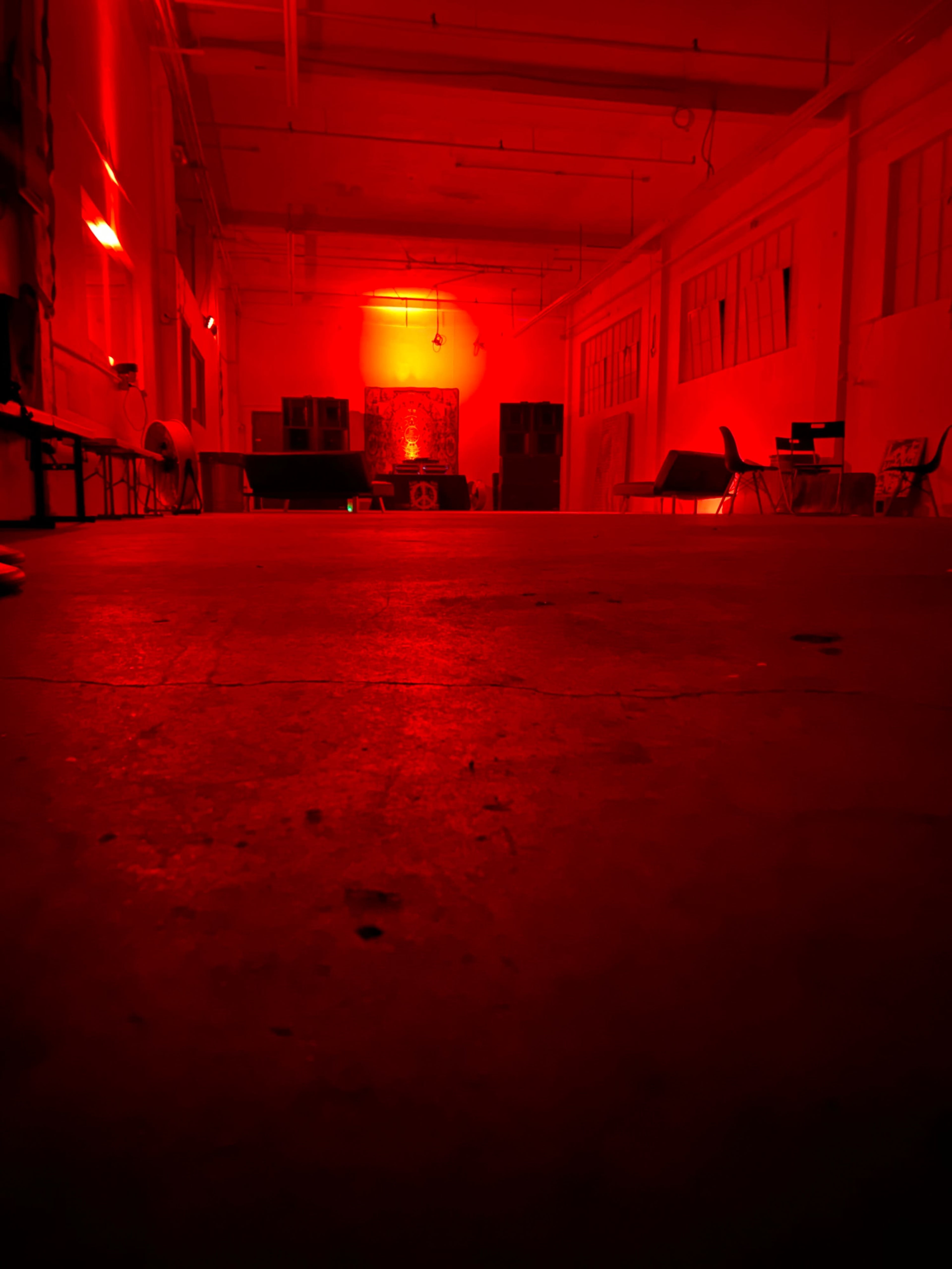 The image shows a large, empty room illuminated with red light, featuring bare walls, a concrete floor, and scattered chairs.