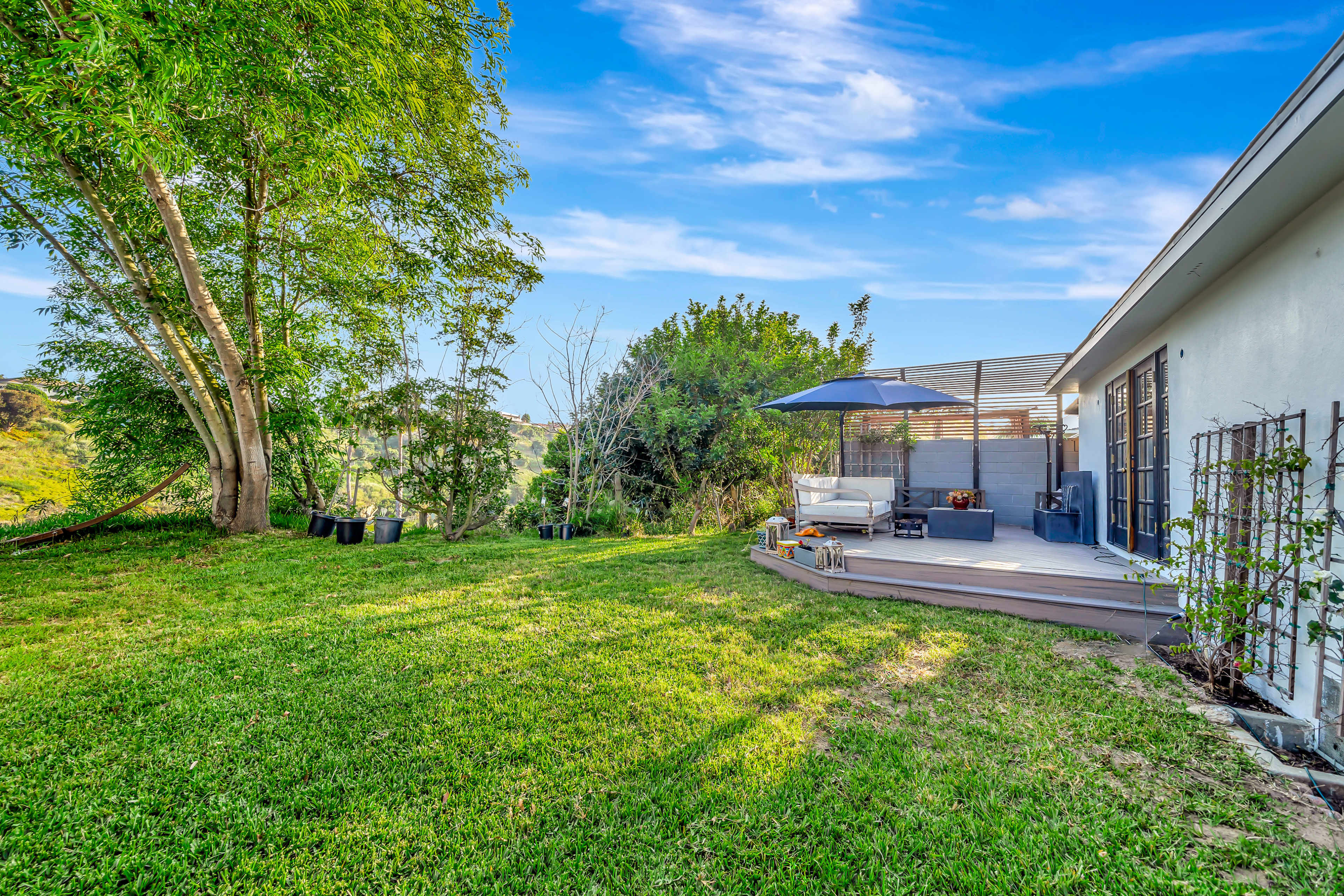 The scene features a backyard with a well-maintained lawn, a patio with outdoor furniture, and several trees in the background.