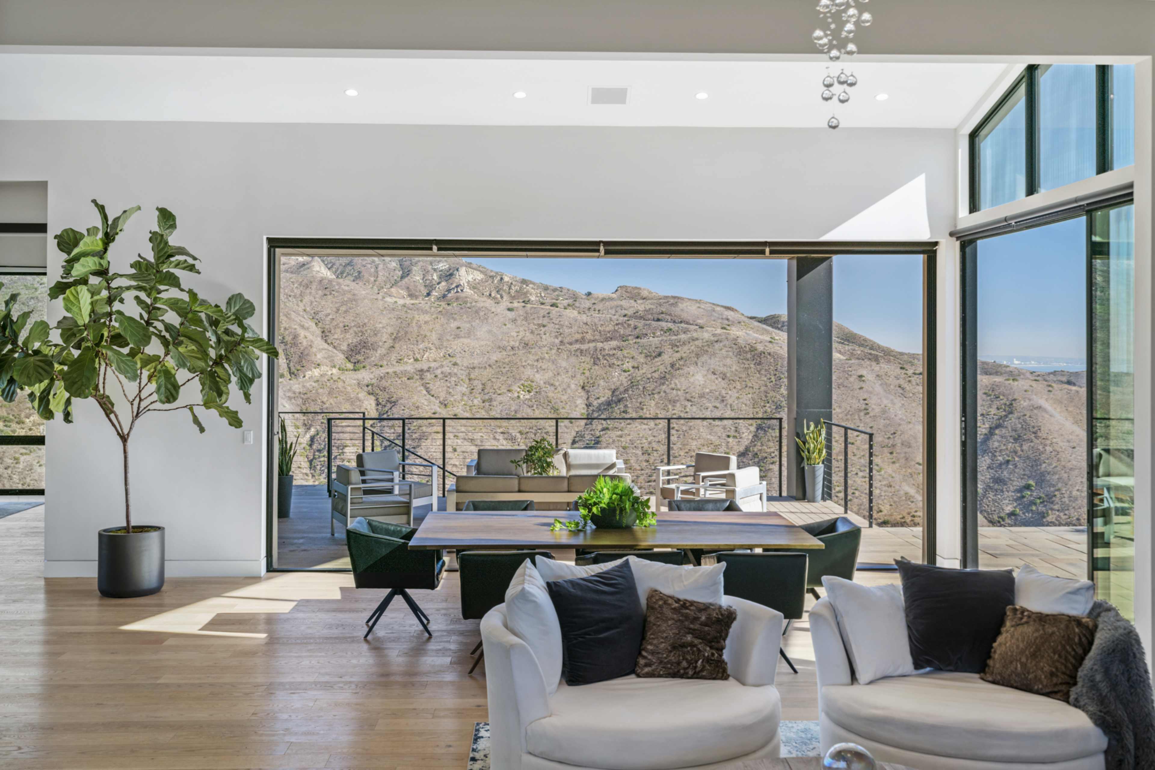A modern living space features large windows revealing a view of rolling hills and a wooden deck furnished with outdoor seating.
