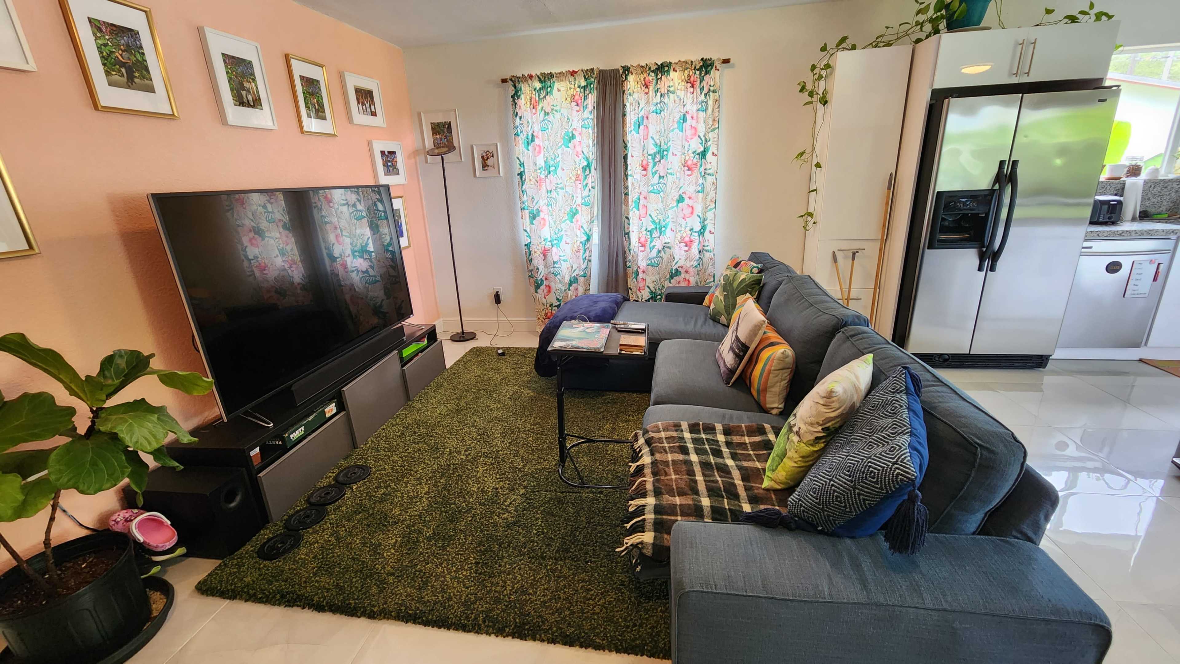 A modern living room features a large sofa with colorful cushions, a TV on a stand, a coffee table, and patterned curtains beside a kitchen area with stainless steel appliances.