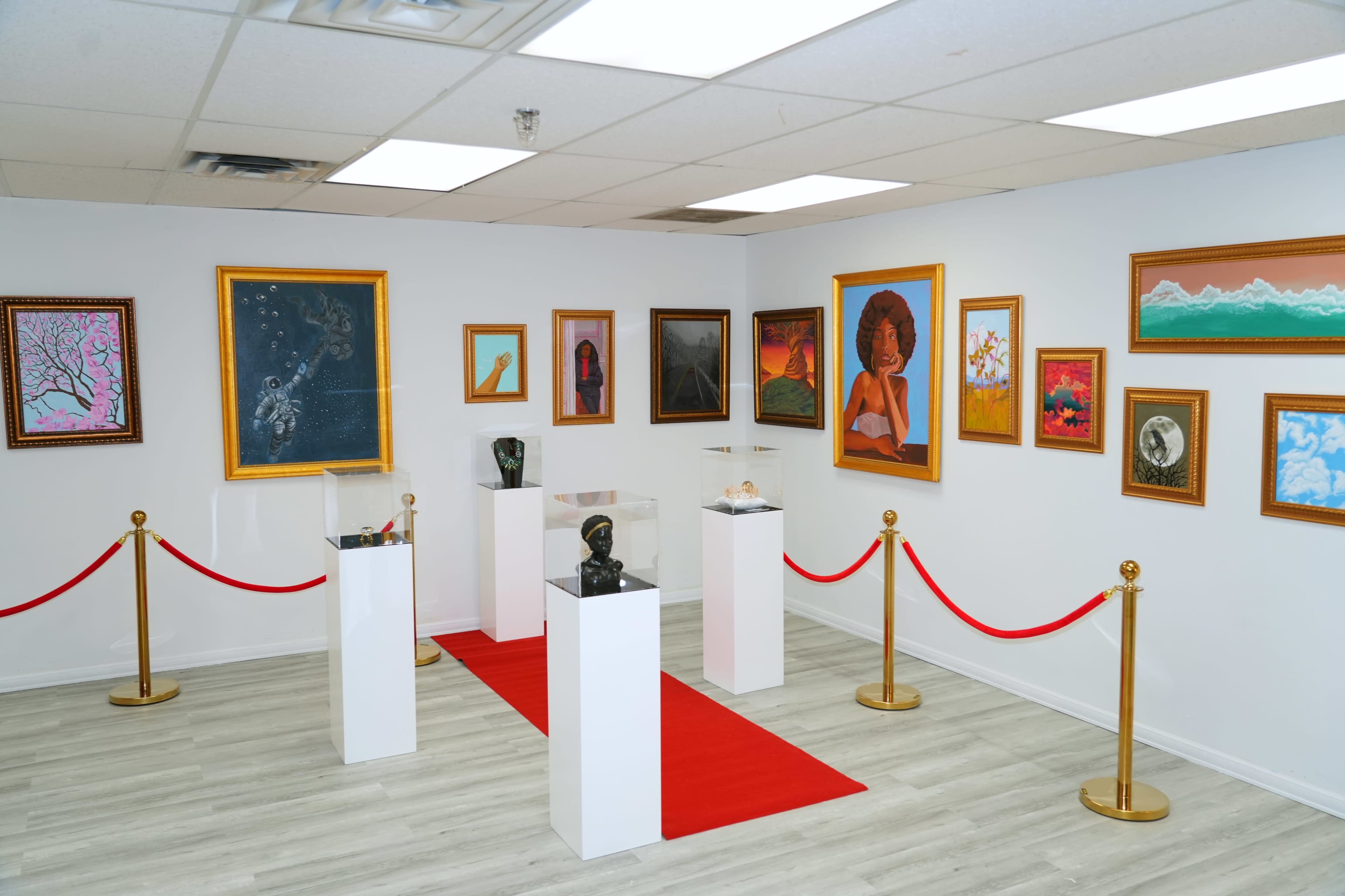 An art gallery featuring several framed paintings on the walls and sculptures displayed on pedestals, with a red carpet and stanchions creating a defined exhibition space.