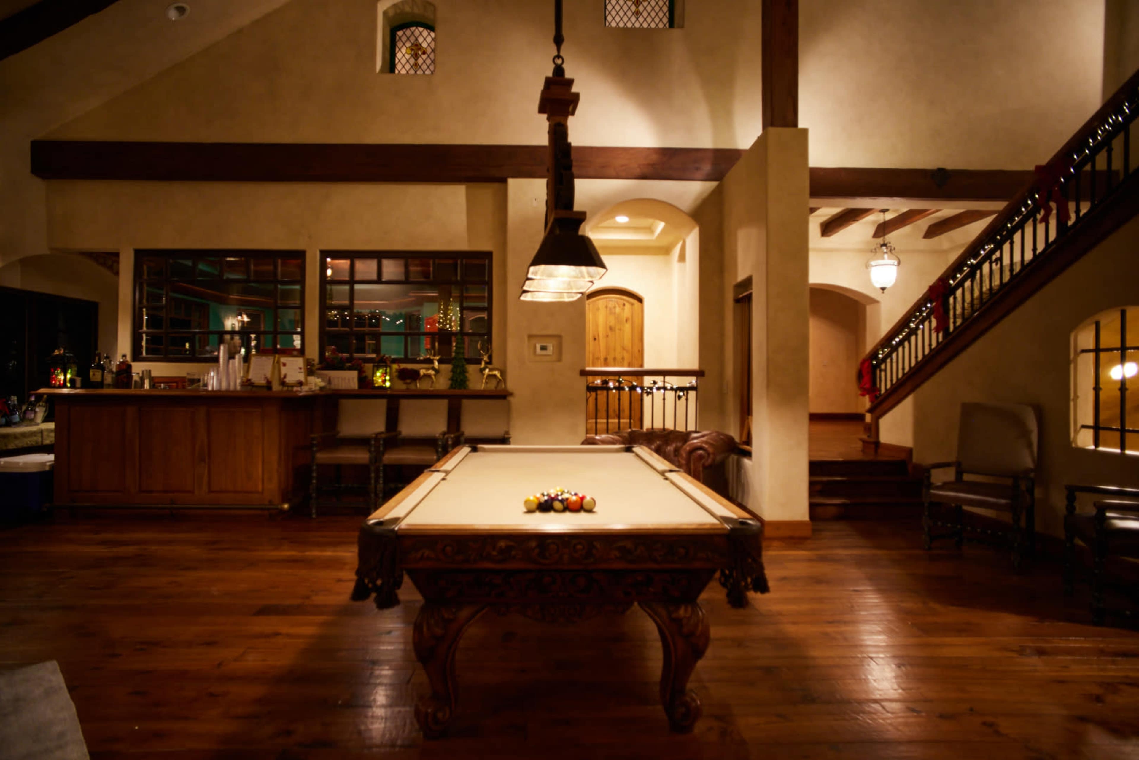 A spacious room with a pool table, bar area, and staircase, featuring wooden beams and warm lighting.