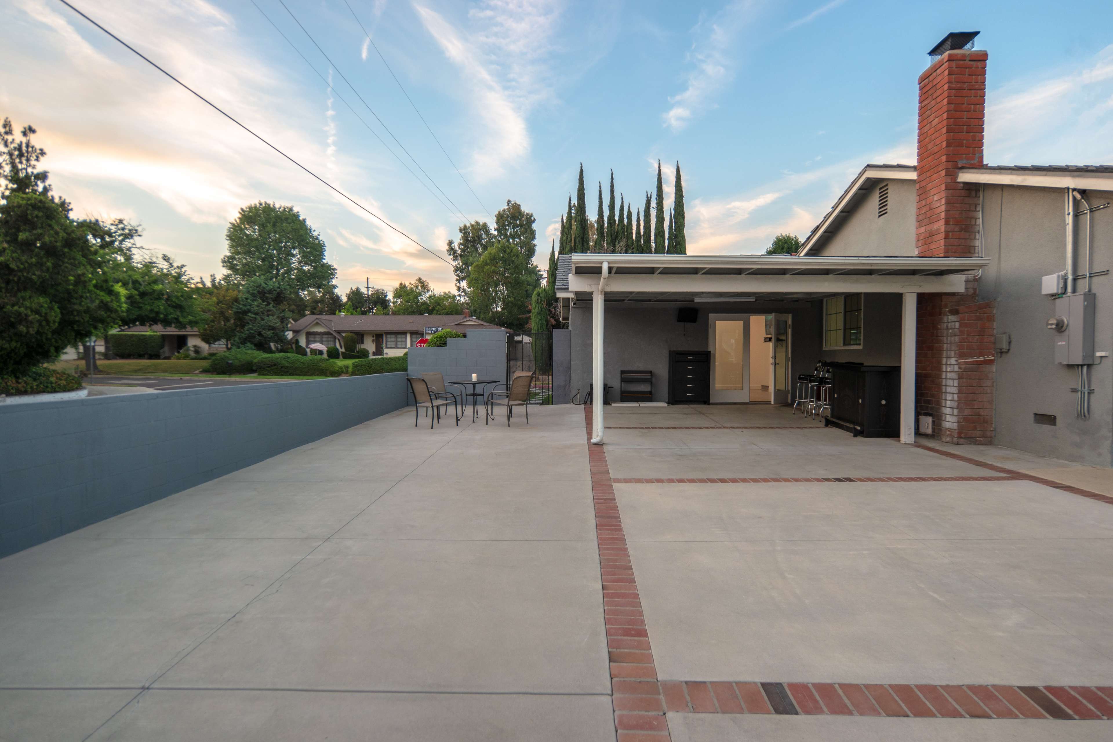 An outdoor patio with a concrete surface, a chimney, and a table set with chairs, adjacent to a house and surrounded by trees.