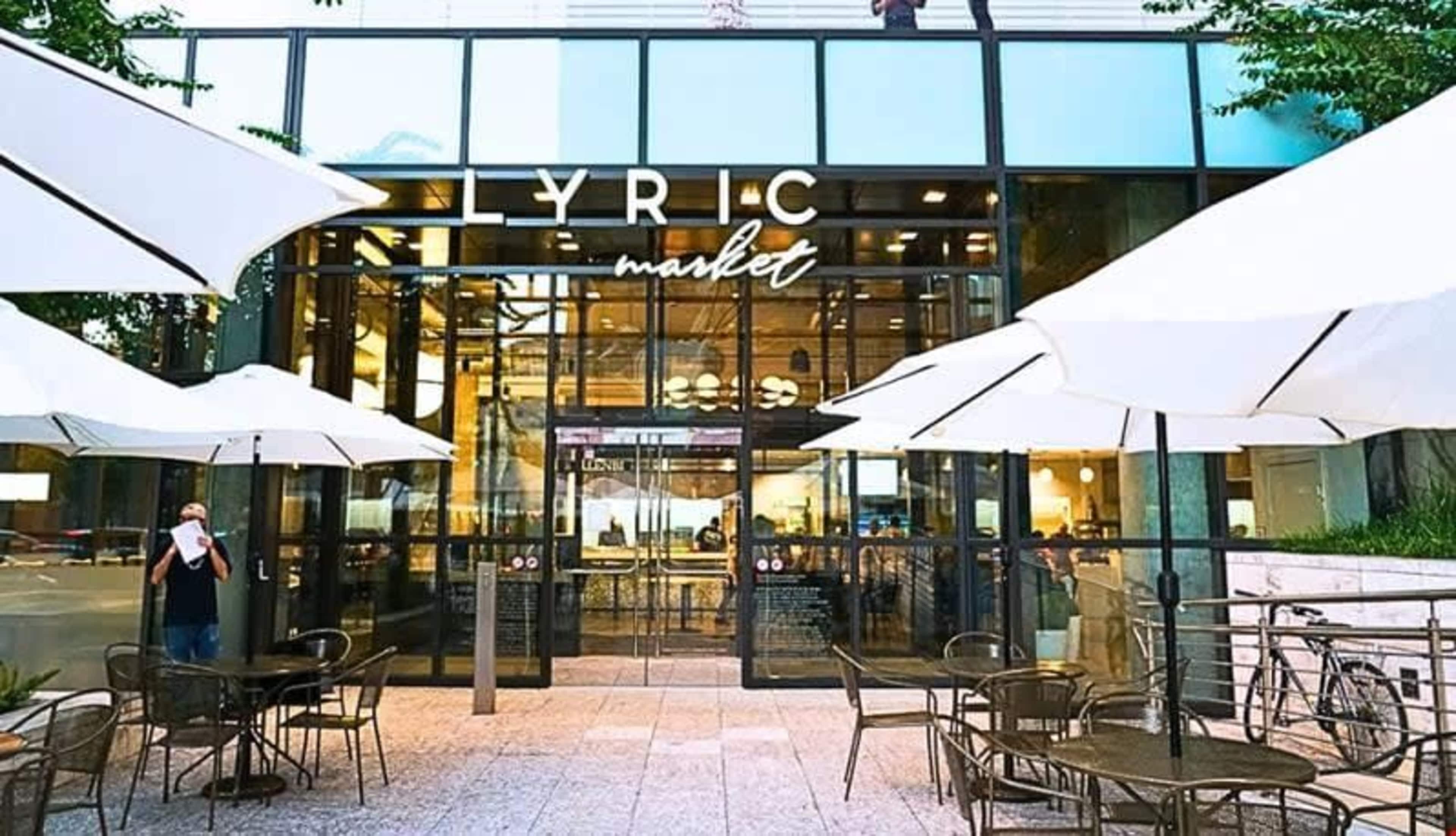 The image shows the entrance of Lyric Market, which features large glass doors and outdoor seating with white umbrellas.