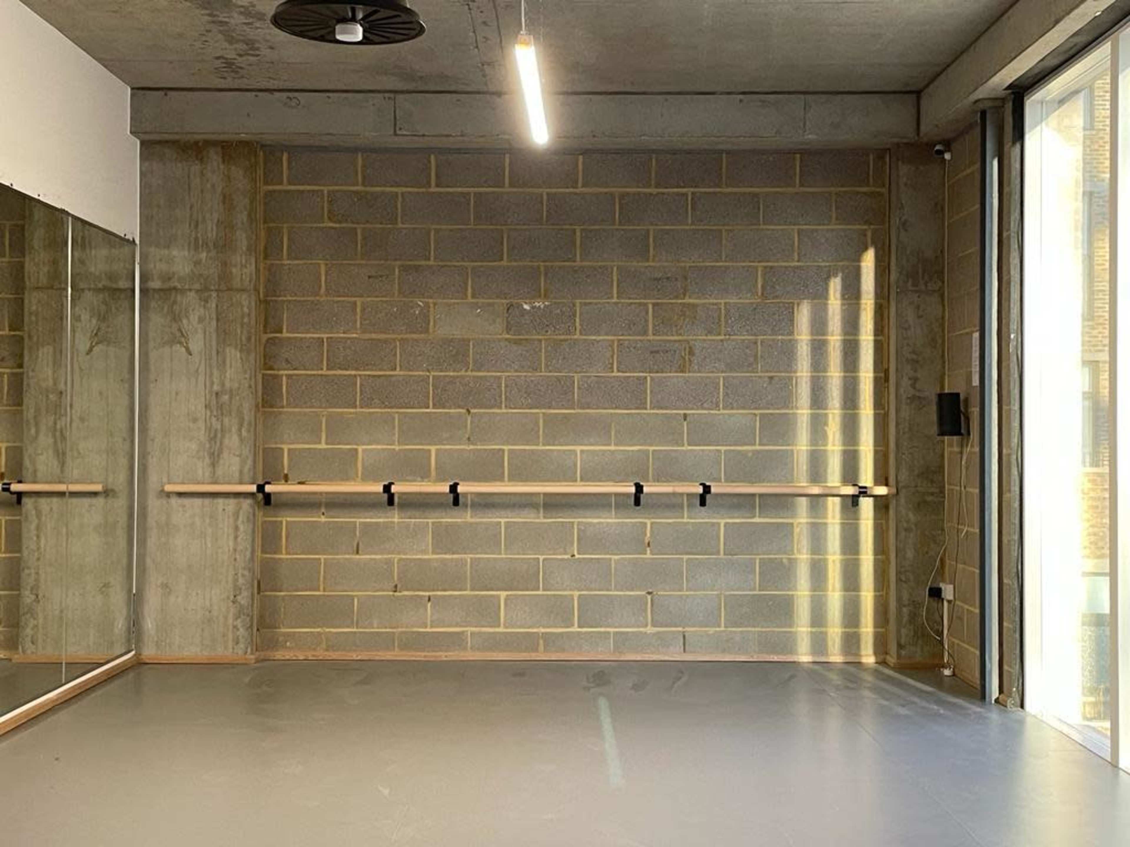 A dance studio with a concrete block wall, a horizontal ballet bar, and large windows allowing natural light to enter.