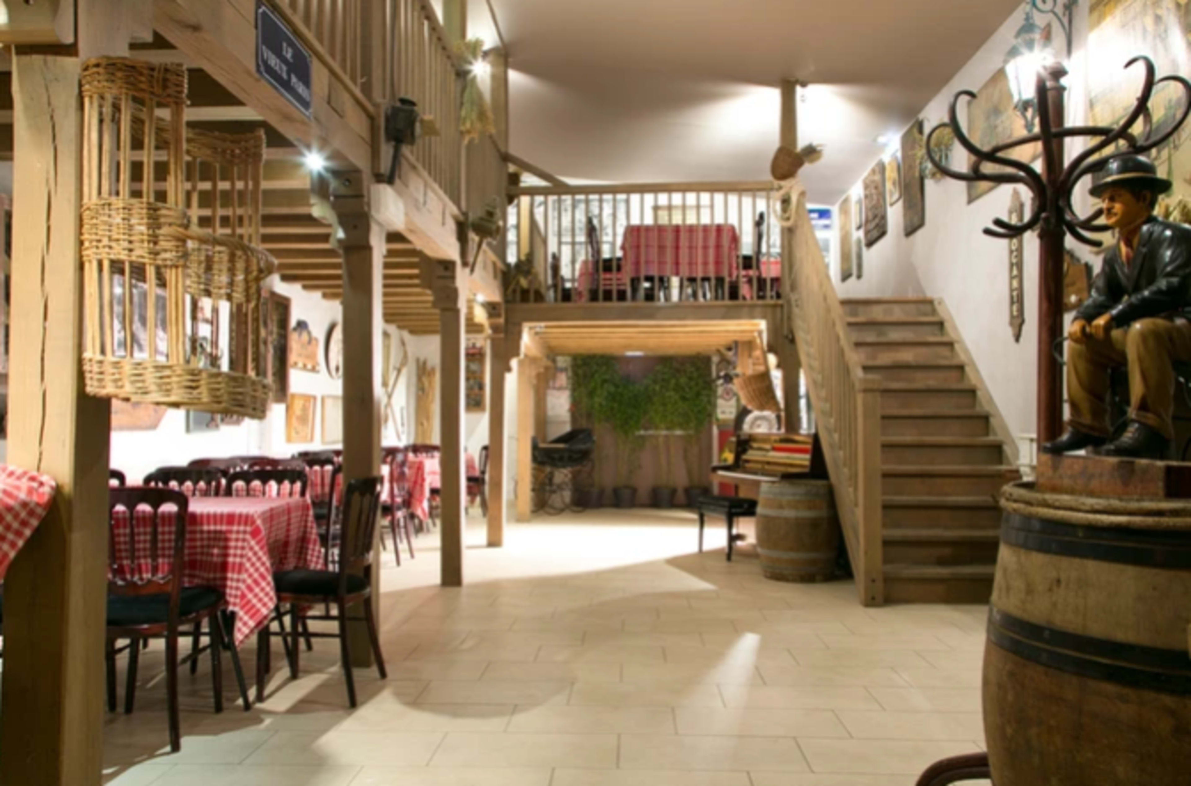 The interior of a rustic restaurant featuring wooden beams, tables with checkered tablecloths, and a staircase leading to an upper level.