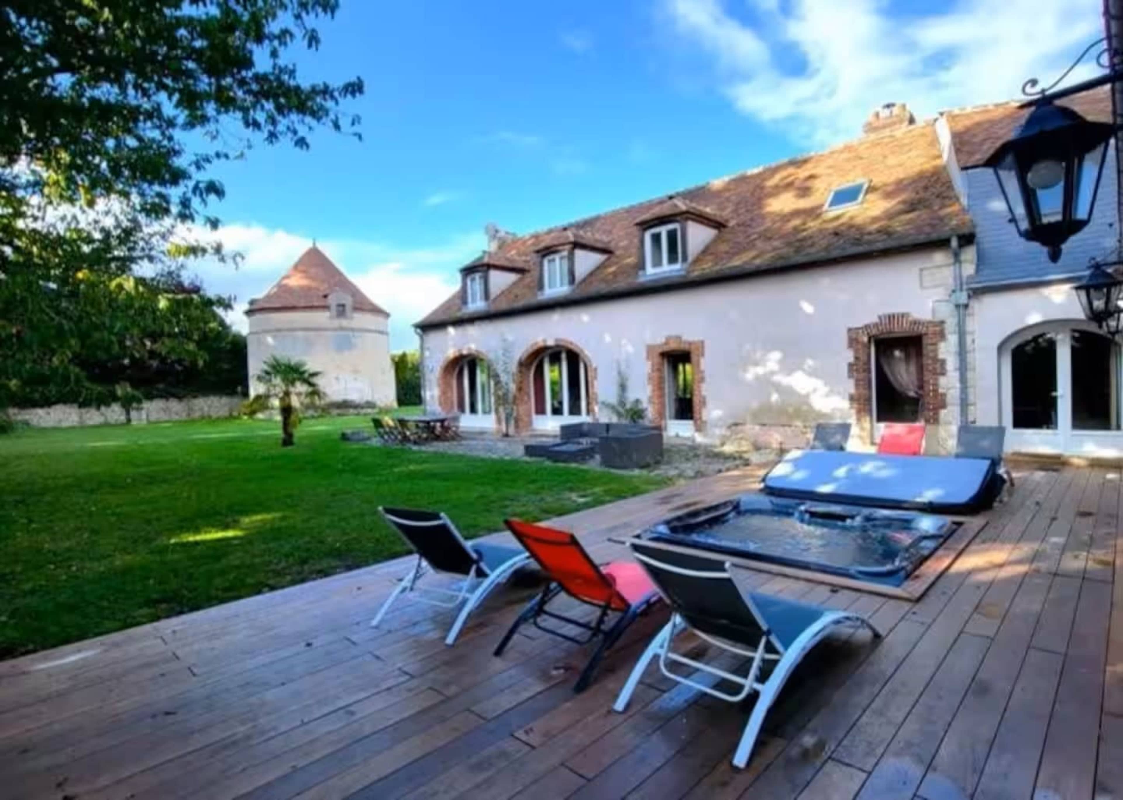 A large house with multiple windows and a wooden deck features lounge chairs and a hot tub, set in a grassy area with trees in the background.