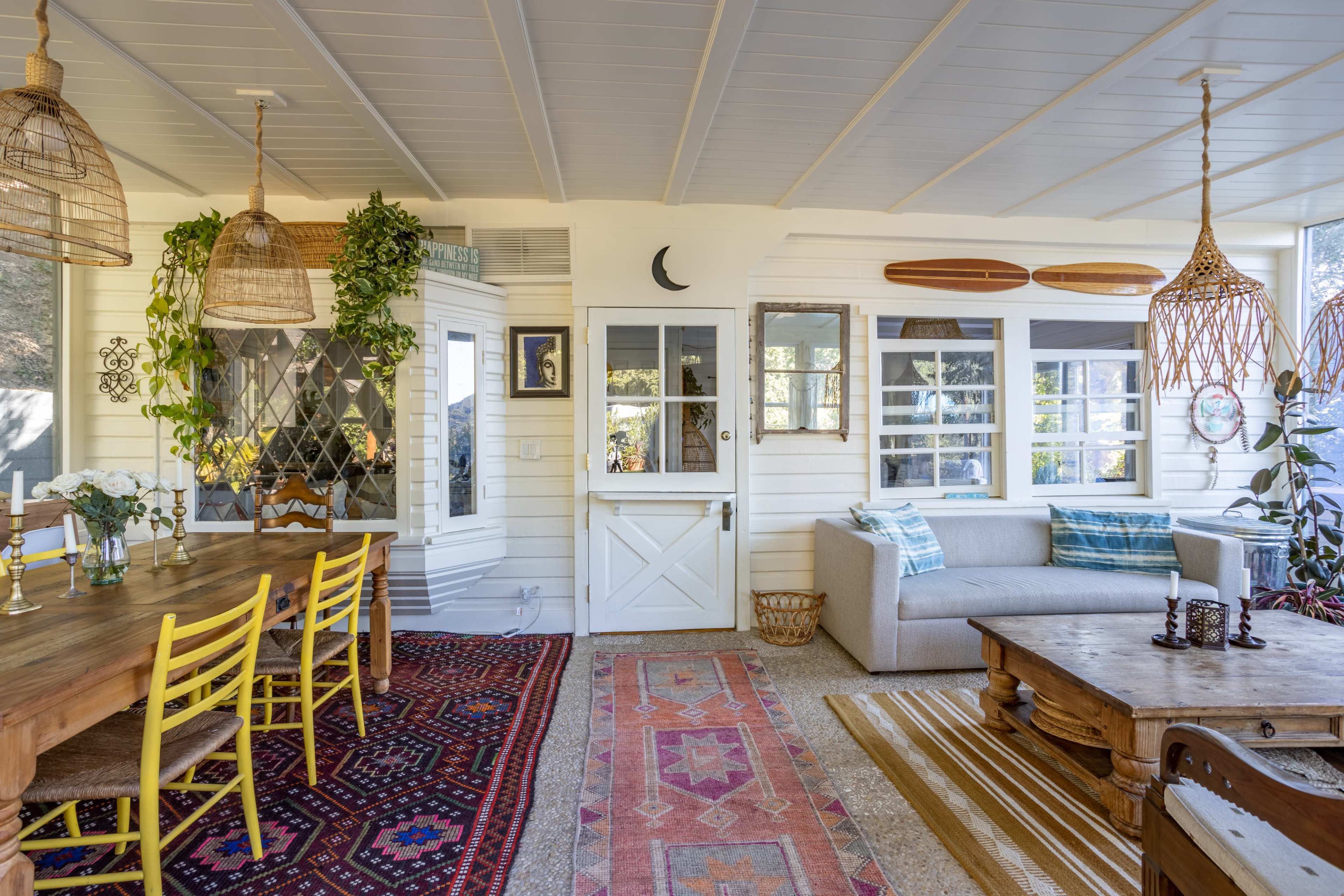 A cozy sunroom with a dining table, bright yellow chairs, a sofa, patterned rugs, and windows adorned with plants.