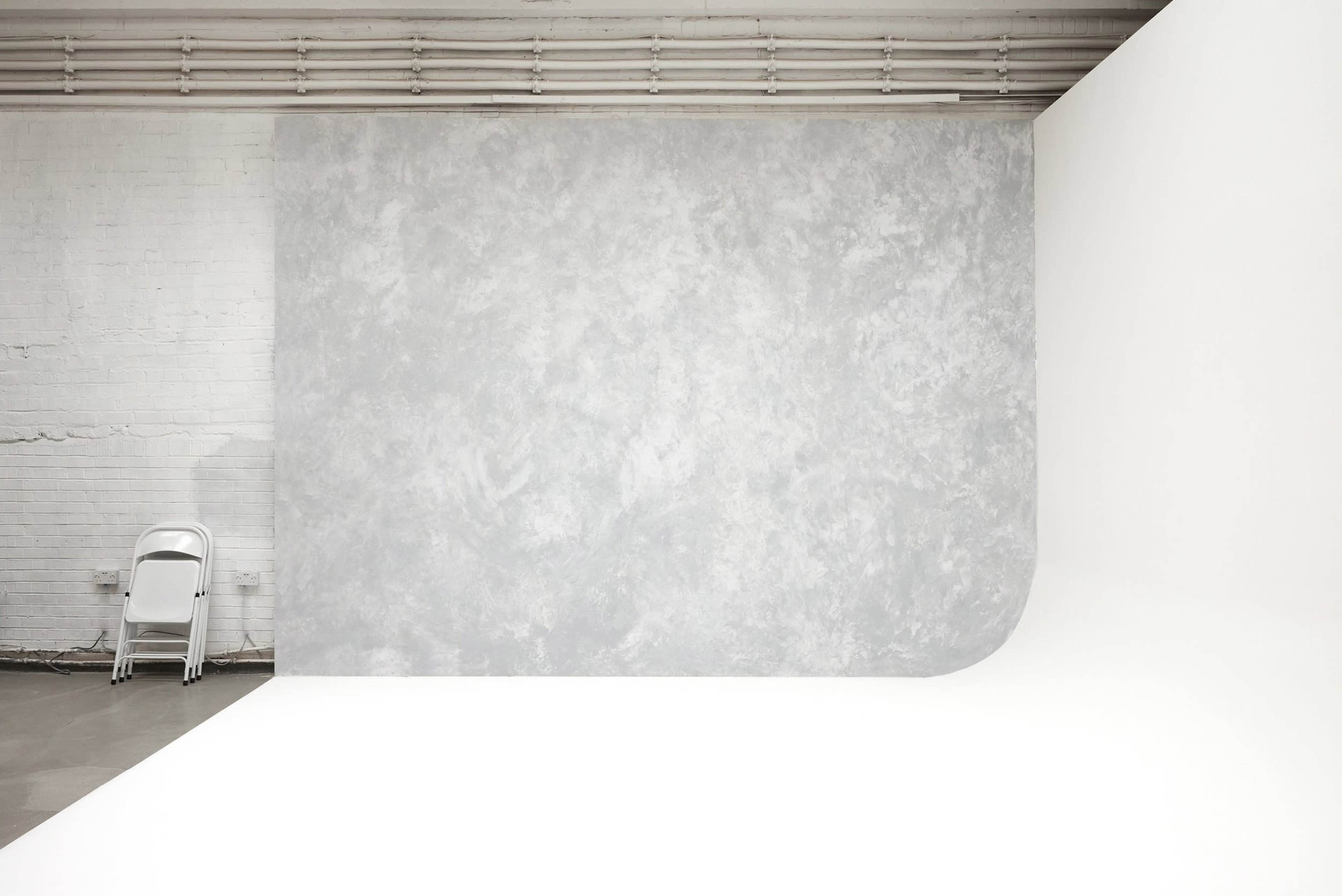 A photography studio with a large, textured gray backdrop and a section of smooth white flooring, alongside a white folding chair against a wall.