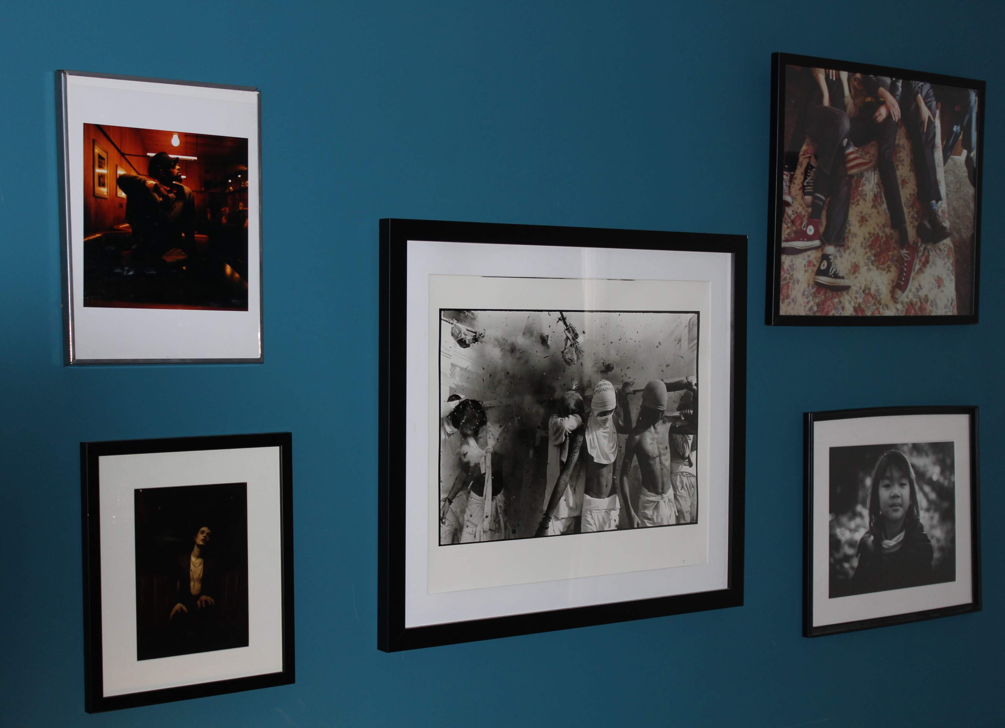 A wall painted in teal with five framed photographs in various styles and sizes arranged on it.