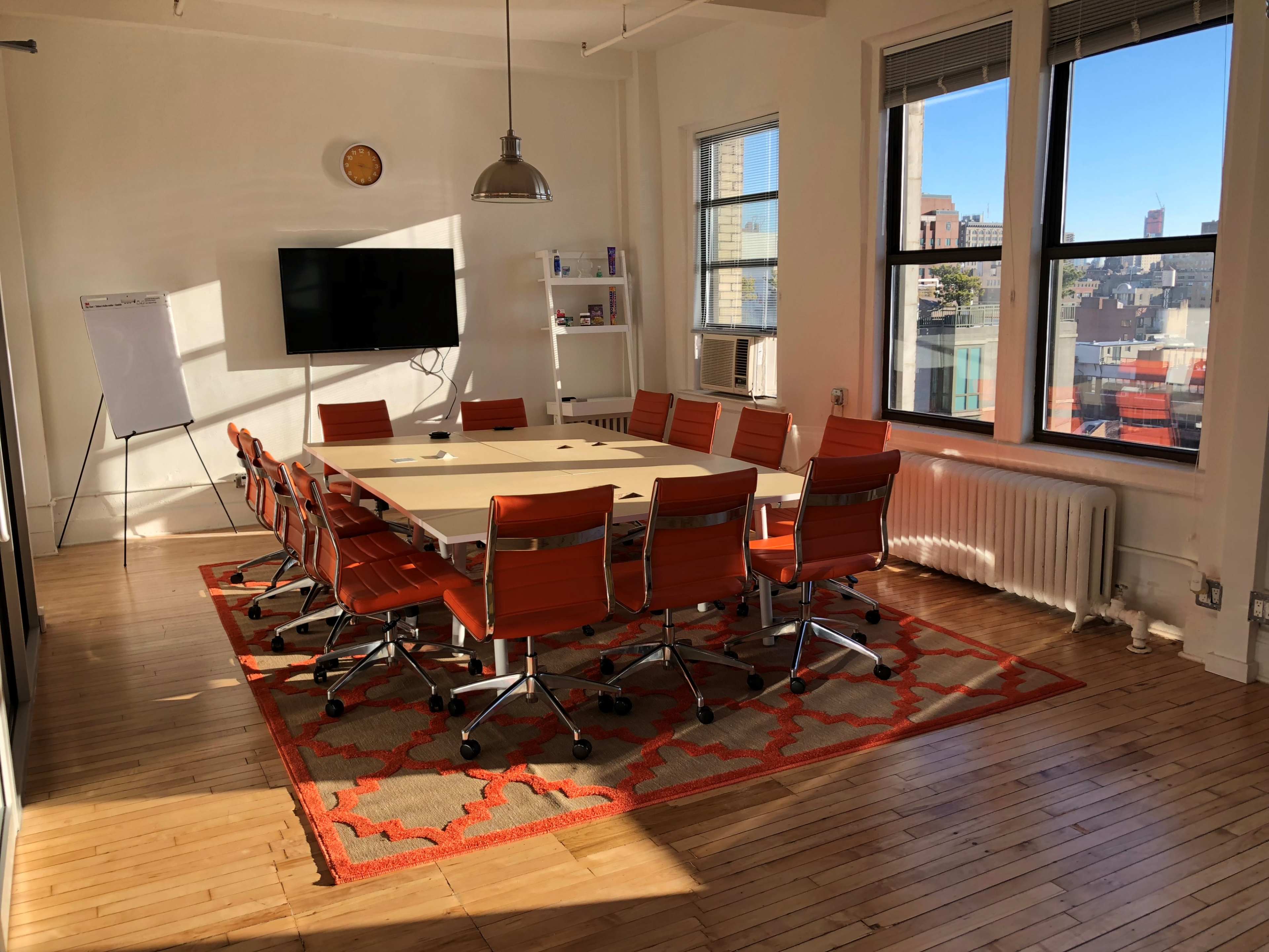 A conference room features a large table surrounded by orange chairs, with a whiteboard and a television visible against the wall, illuminated by natural light from large windows.
