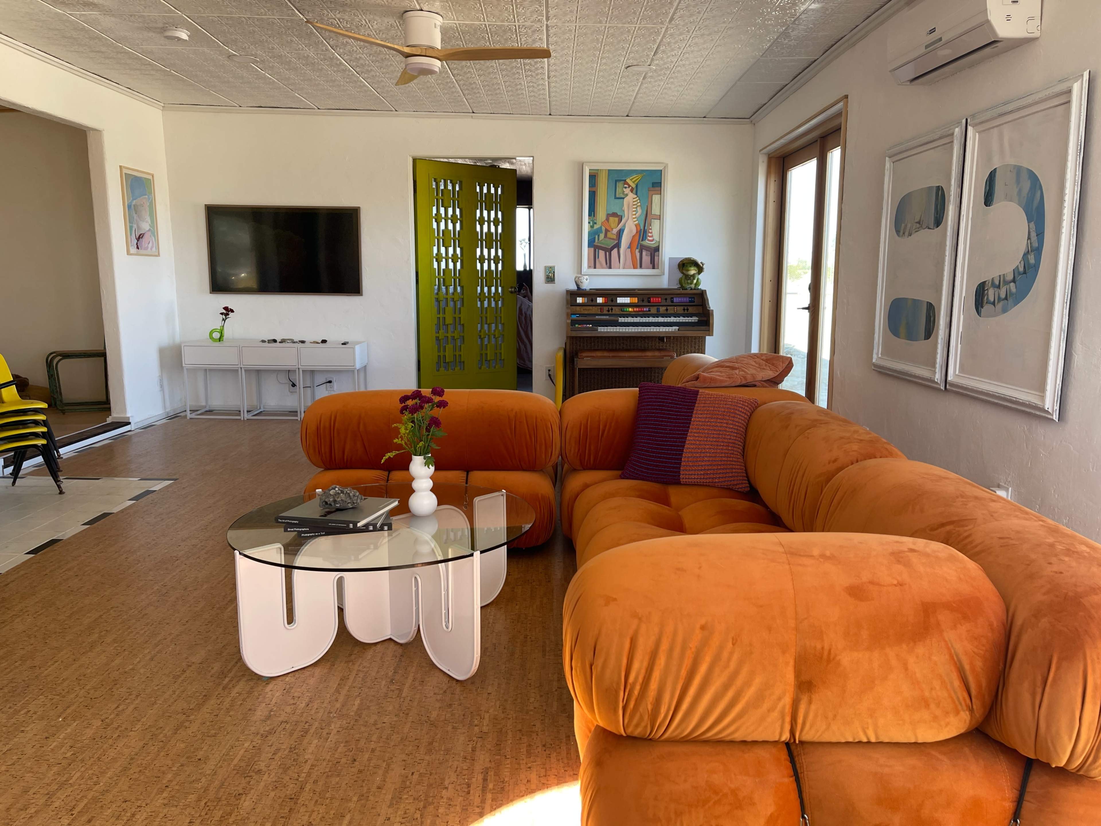 The room features an orange sectional couch, a glass coffee table, and colorful artwork, with a television and a vintage piano visible in the background.