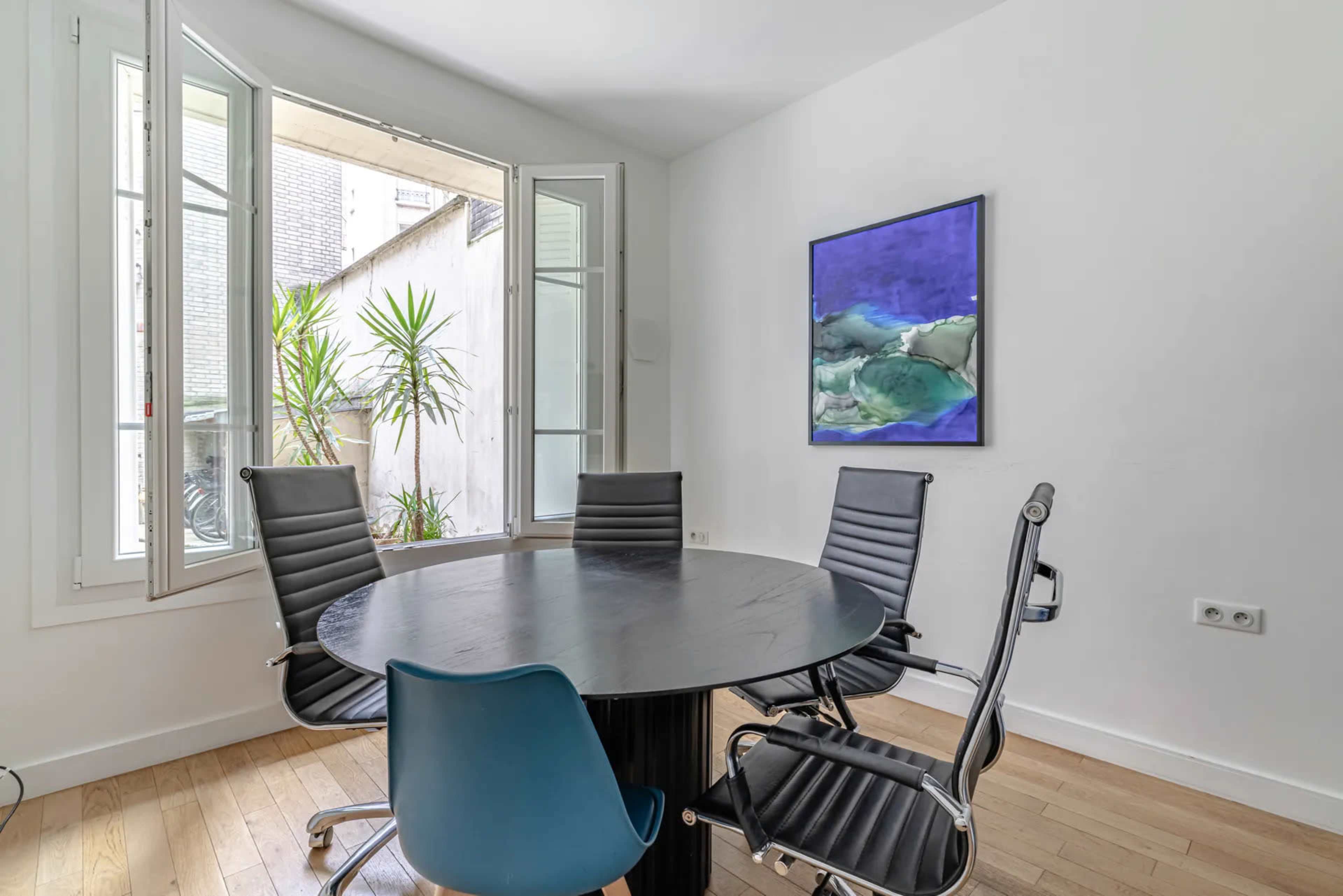 A round black table is surrounded by four chairs in a bright room with large windows and a colorful painting on the wall.