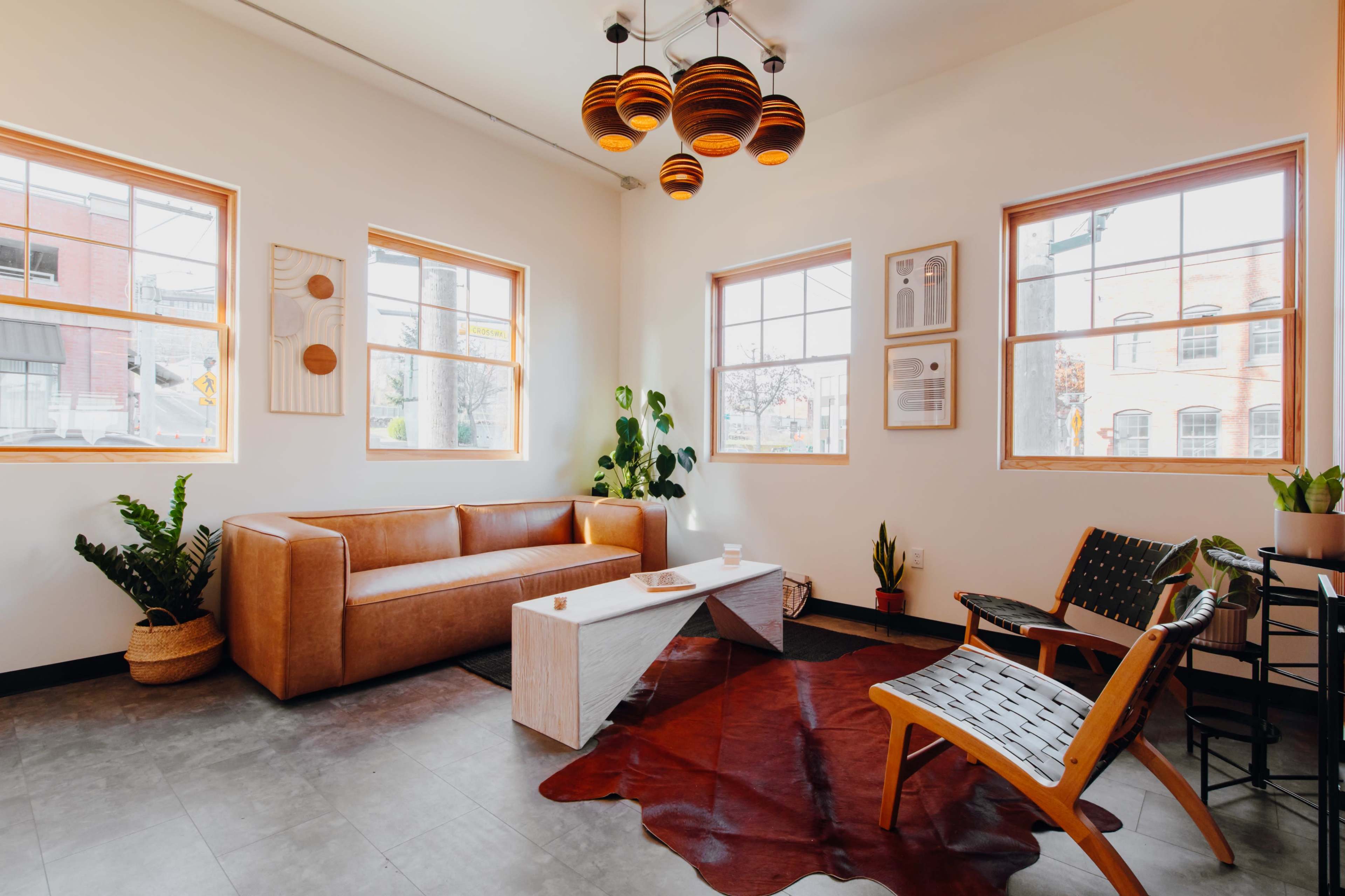 A brightly lit interior featuring a brown leather sofa, a wooden coffee table, and a unique light fixture, complemented by potted plants and large windows.