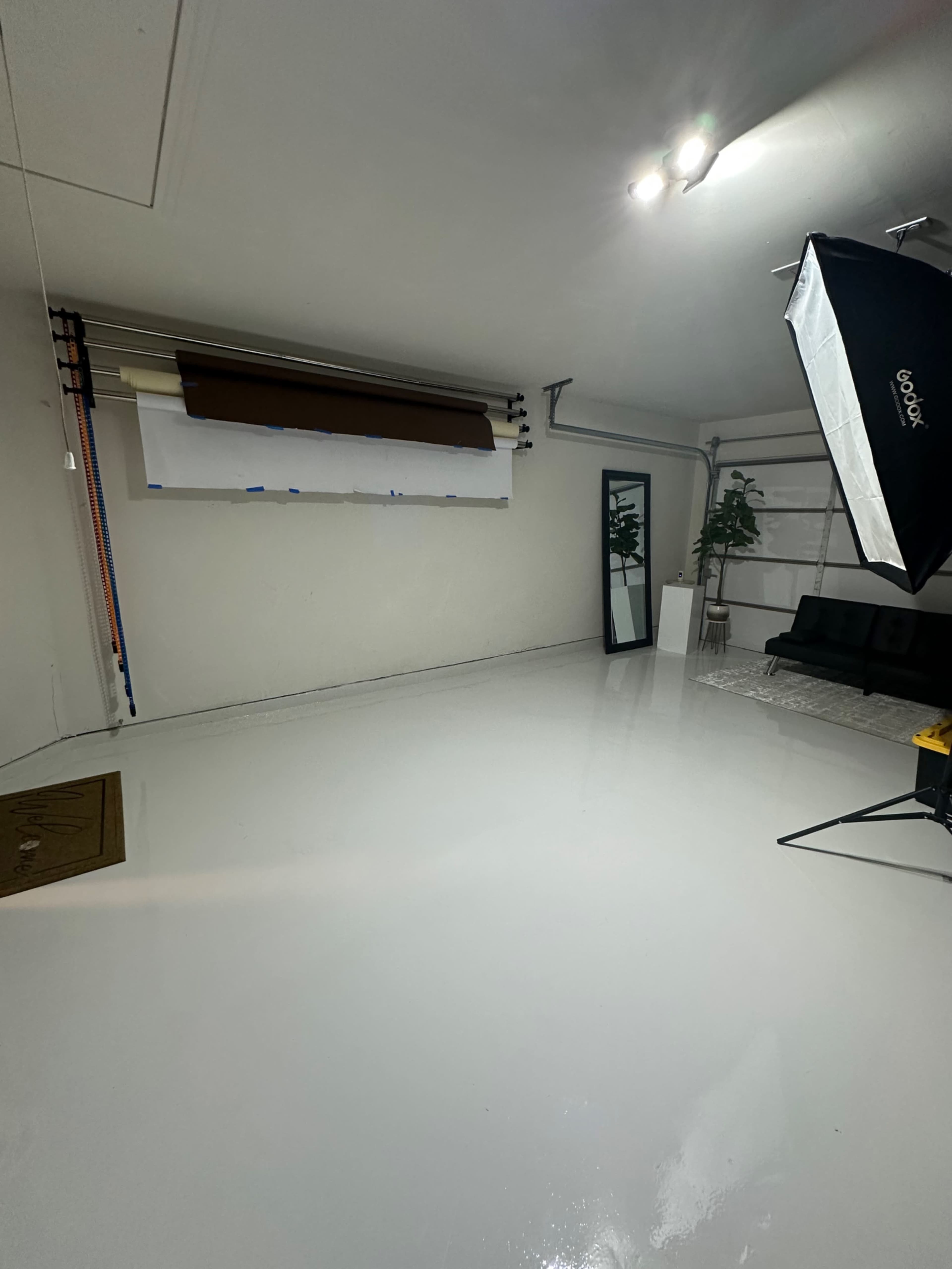 The image shows a clean, well-lit room with a glossy white floor, a large mirror, a black sofa, and a photography softbox setup in one corner.
