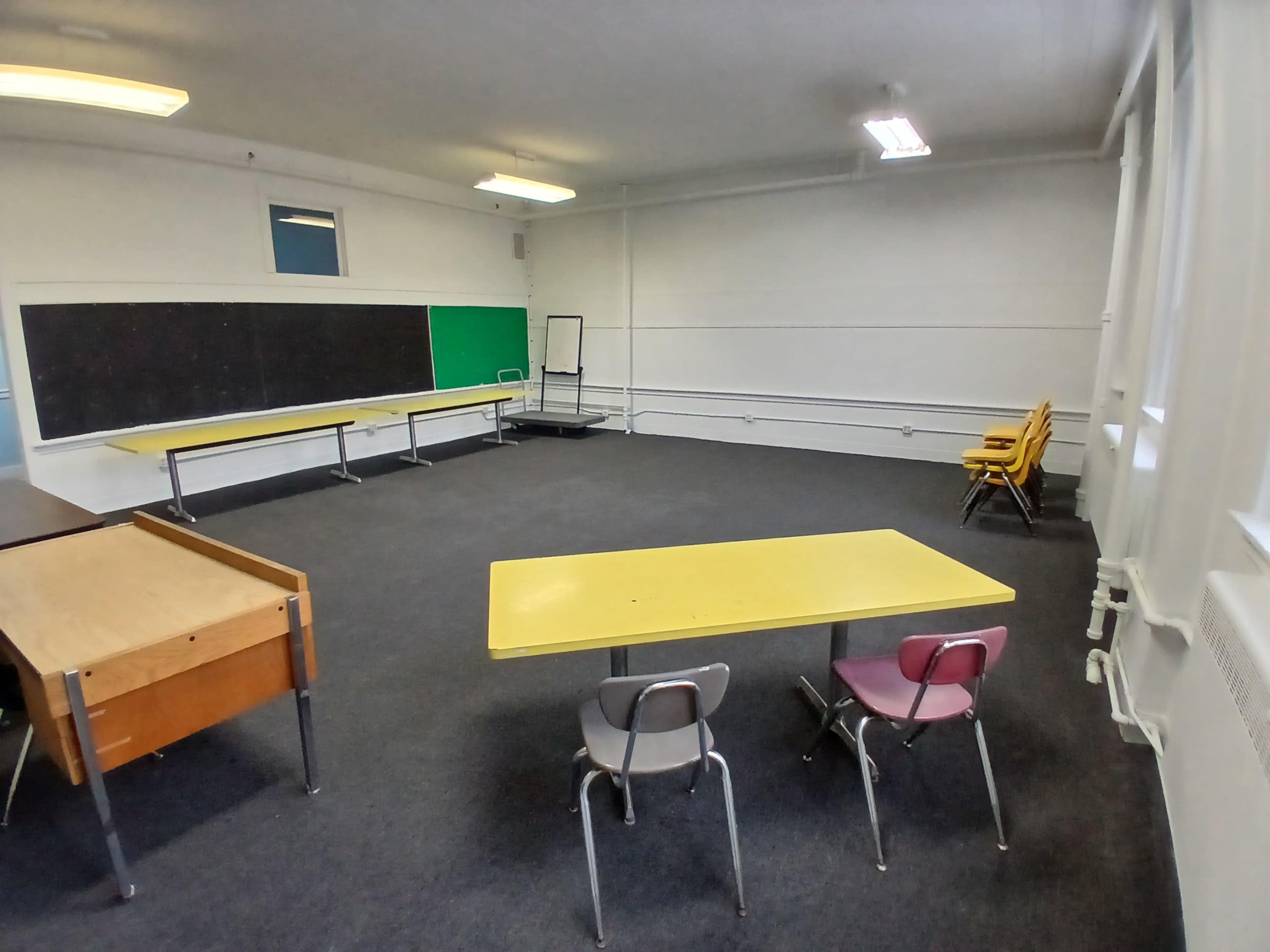 The image shows a classroom with yellow tables, a chalkboard, and a few stacks of wooden chairs against the wall.