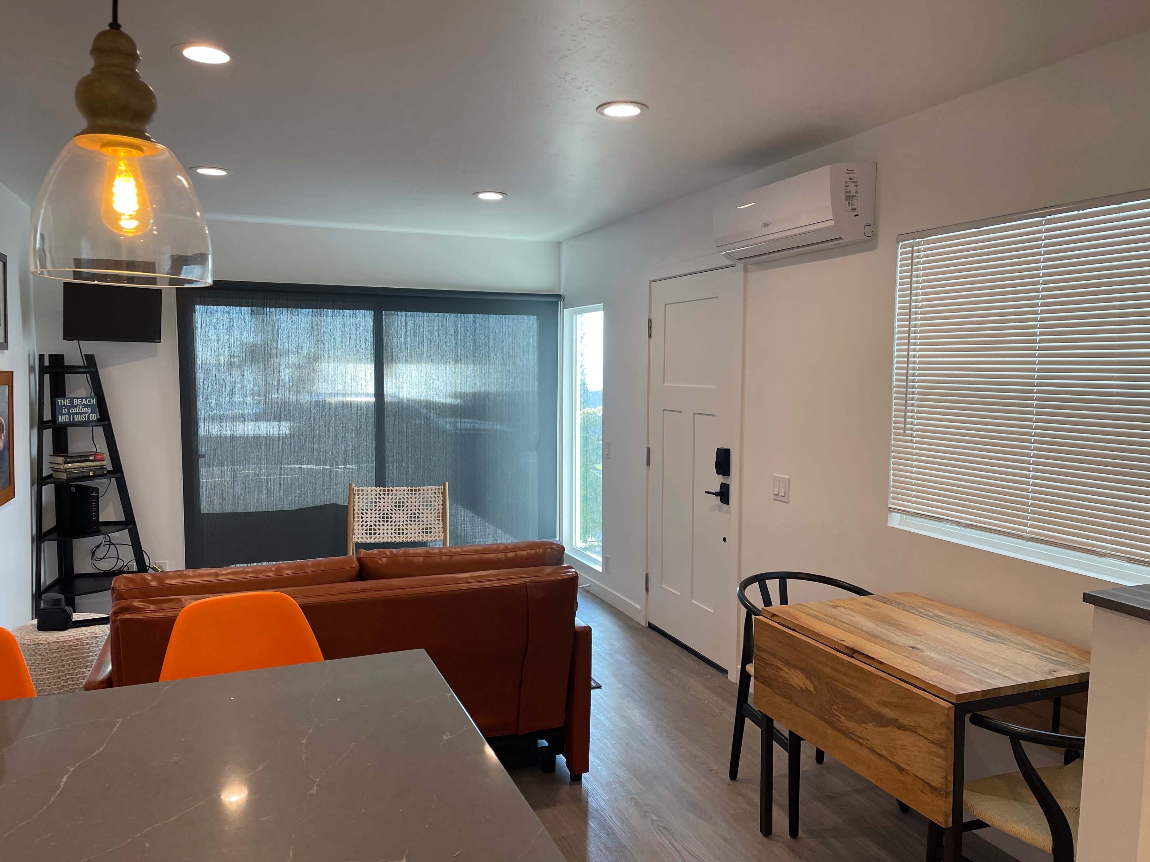 The image shows a modern living space featuring a brown sofa, a wooden dining table with black chairs, and a large window with a view.