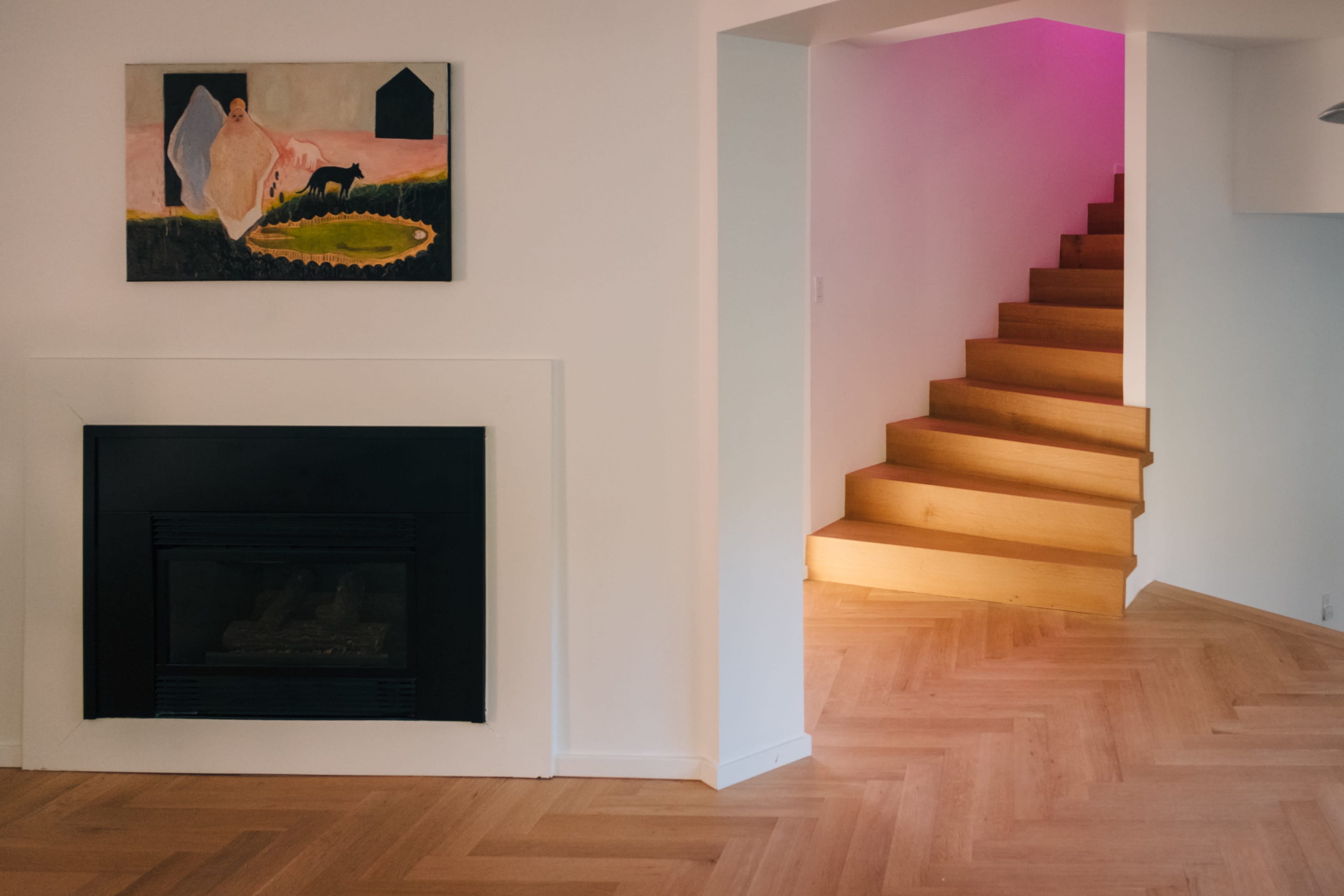 The image shows a modern interior featuring a fireplace, a painting on the wall, and a staircase illuminated with pink light.