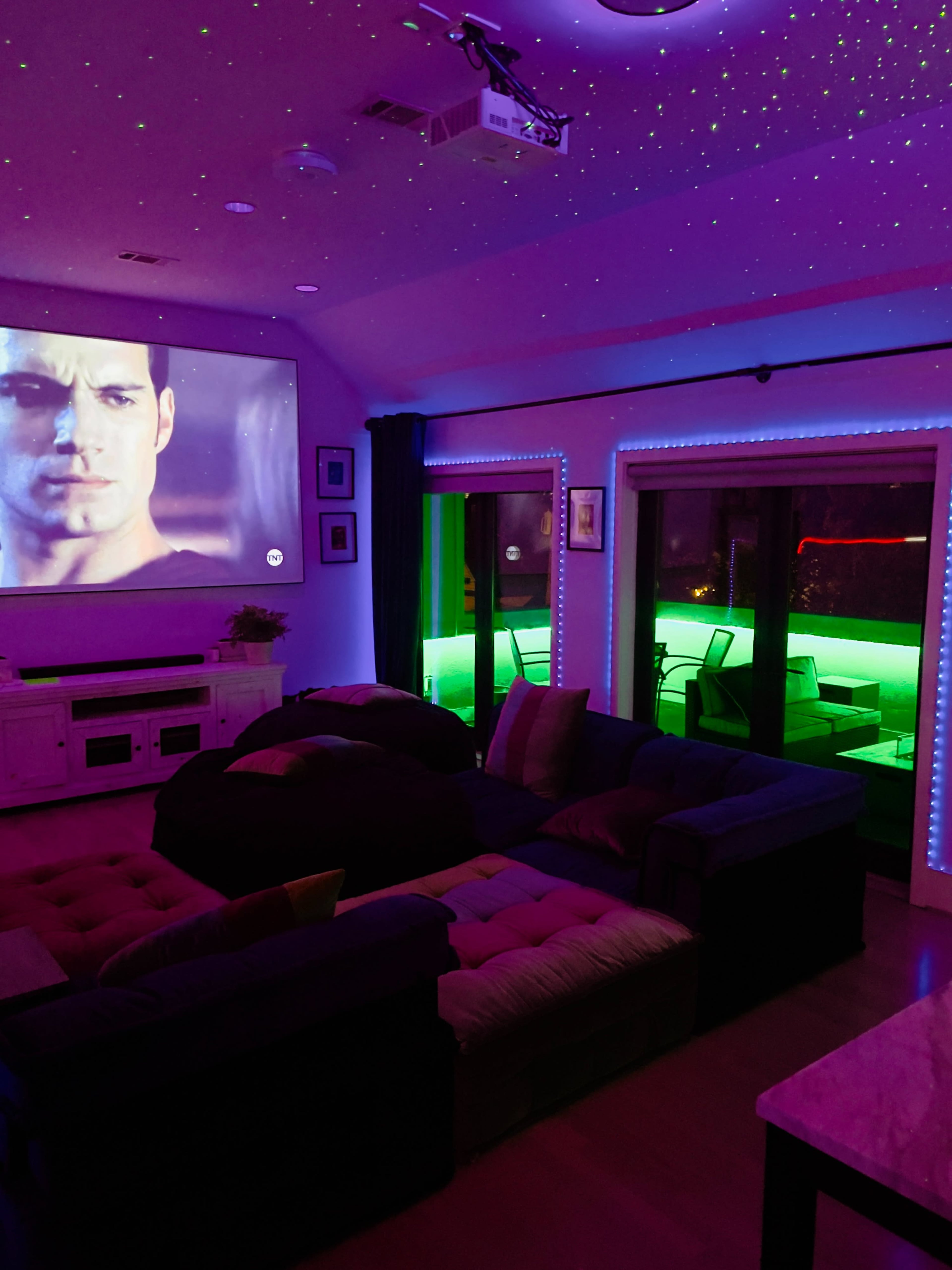 A home theater setup features a large projection screen displaying a movie scene, surrounded by ambient green and blue LED lighting.