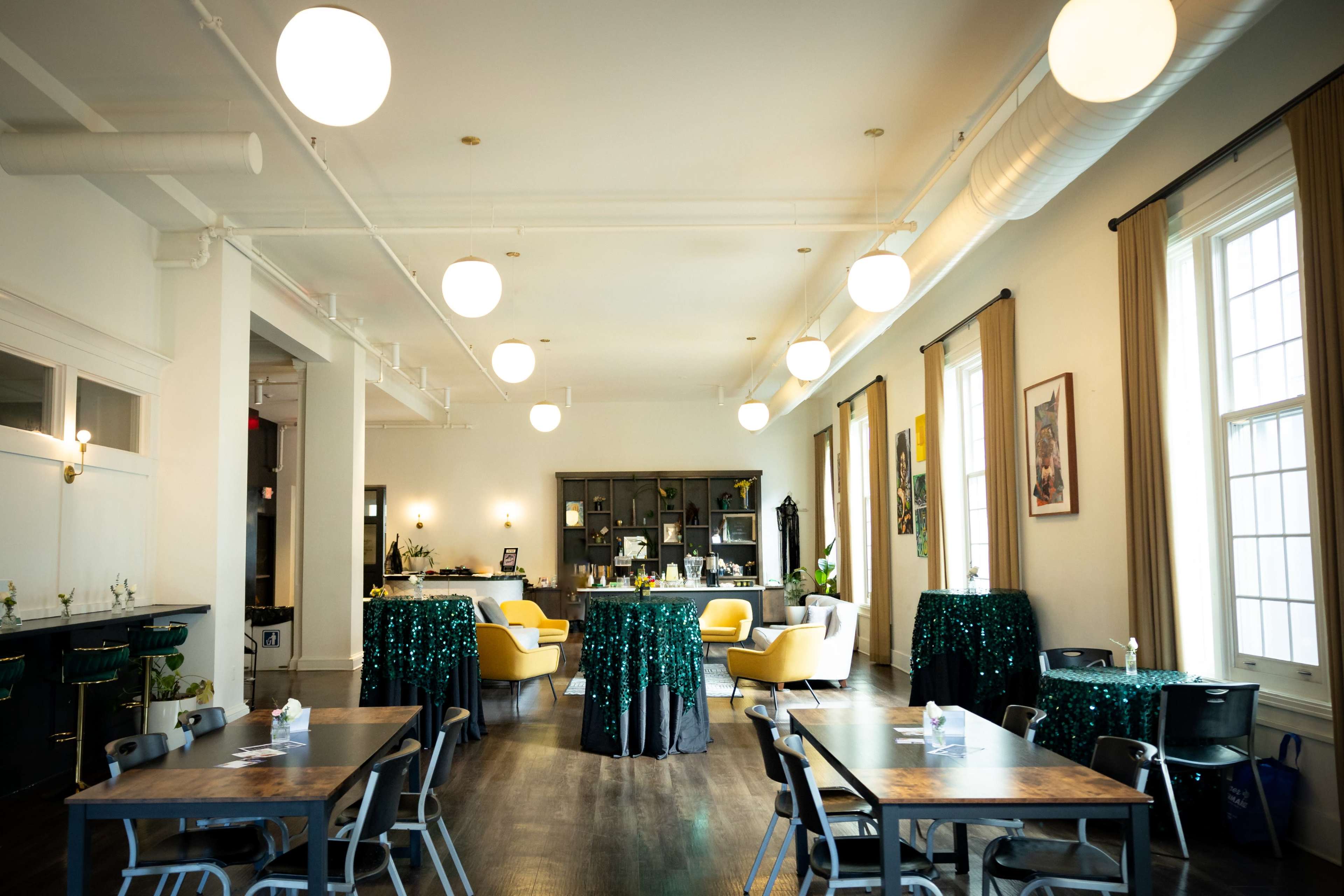 A spacious event space features tables with green sequin tablecloths and a bar area, illuminated by round pendant lights.