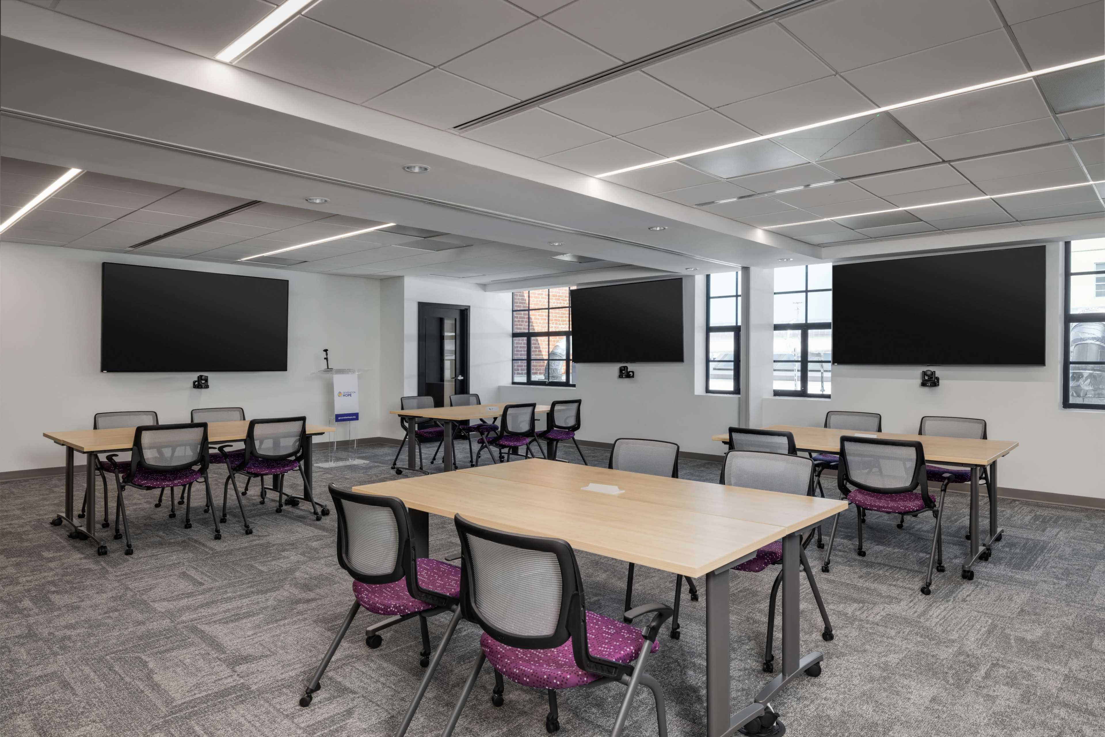 The image shows a spacious classroom setup featuring several tables with chairs, two large screens on the walls, and large windows allowing natural light.
