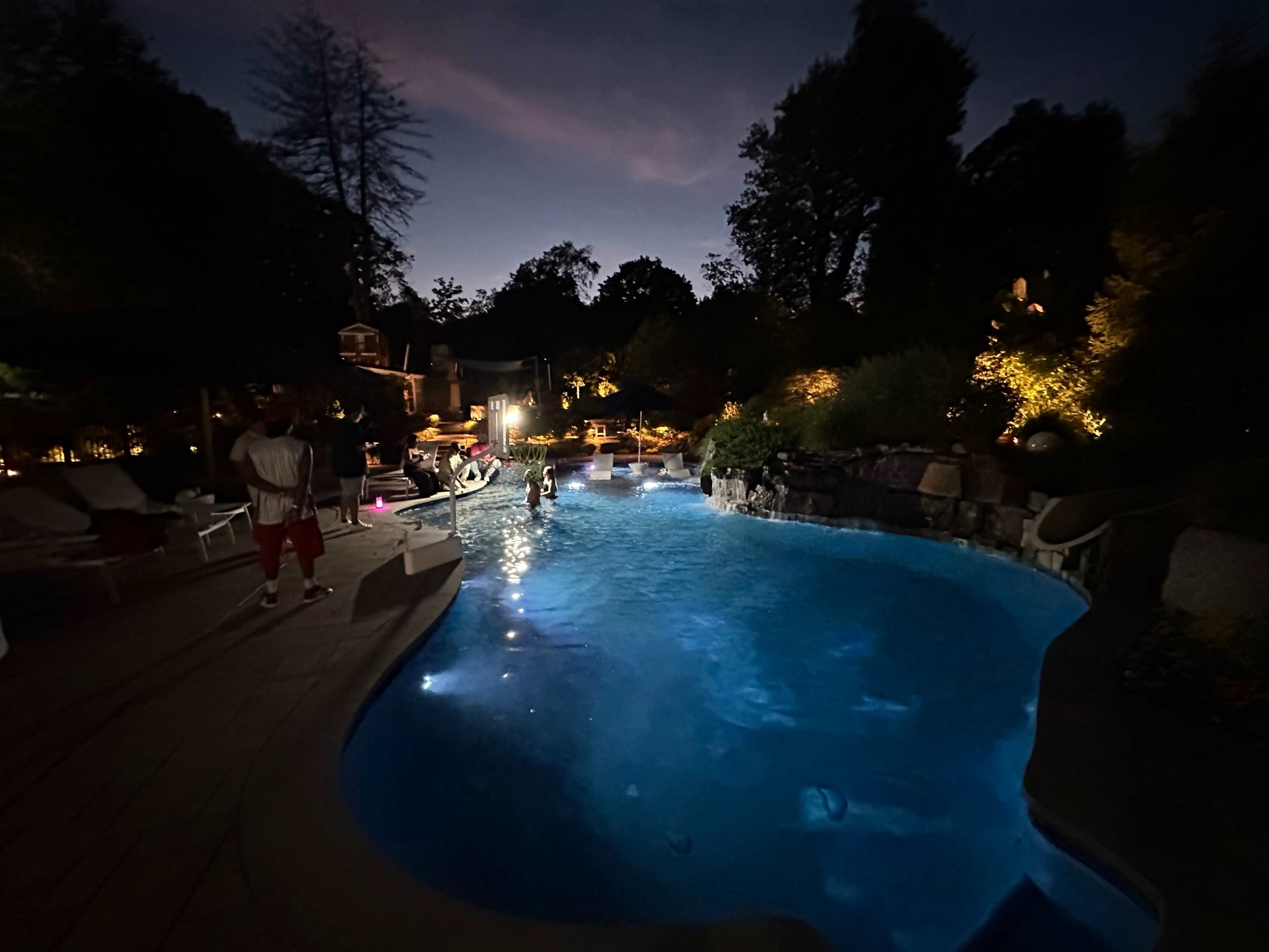 A nighttime scene depicts a swimming pool surrounded by lush landscaping and softly illuminated by ambient lights.
