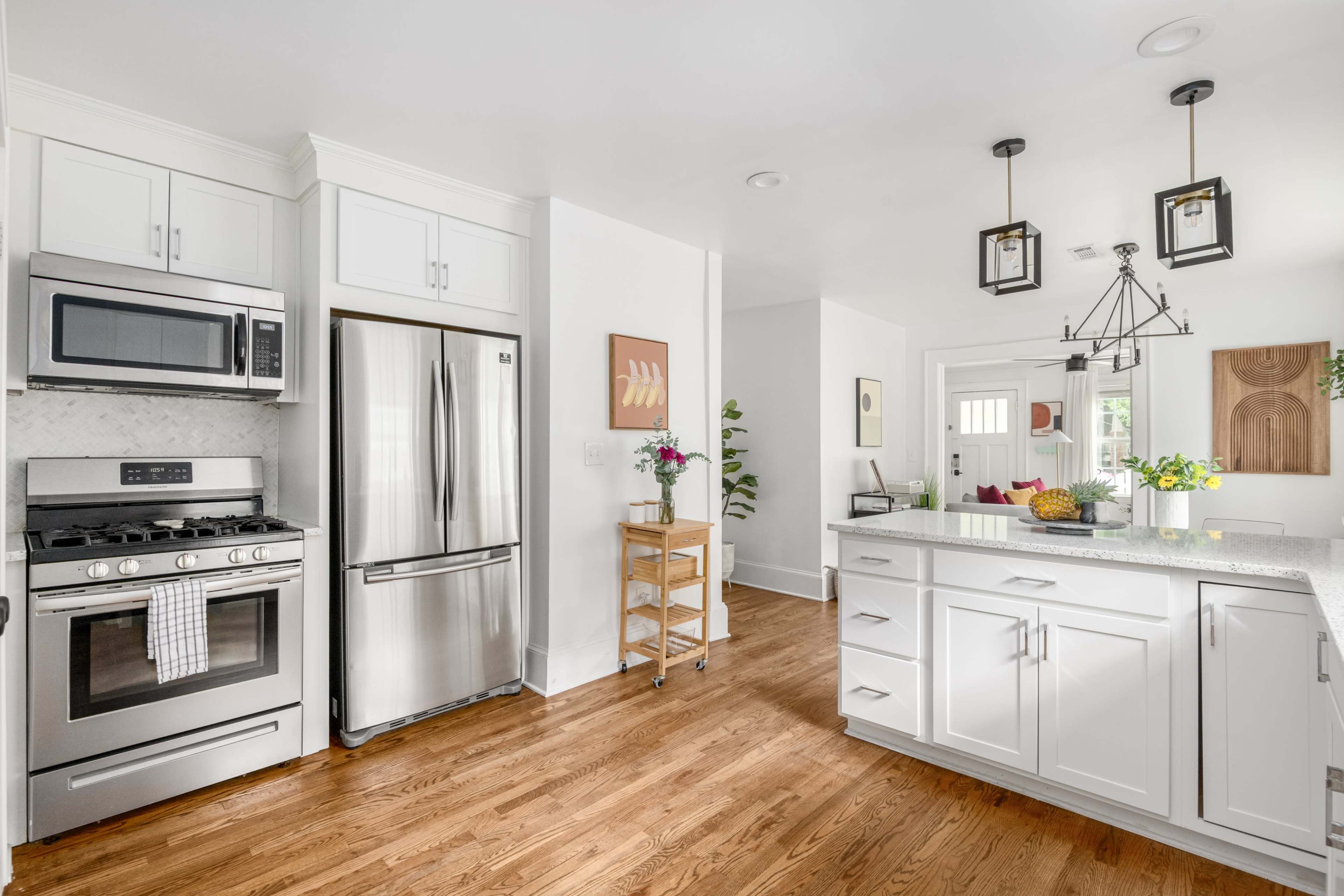 The image shows a modern kitchen with stainless steel appliances, white cabinetry, and a central island with a granite countertop, alongside a living area with a small table and chairs in the background.